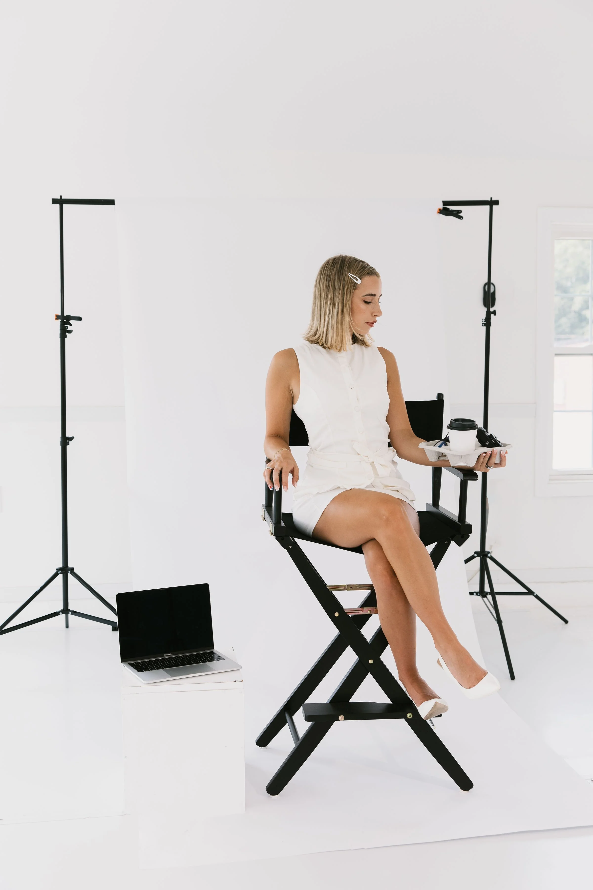 A woman sitting on a director's chair in a bright, minimalist studio with photography equipment, holding a tray with coffee and snacks, with a laptop on a white cube nearby.