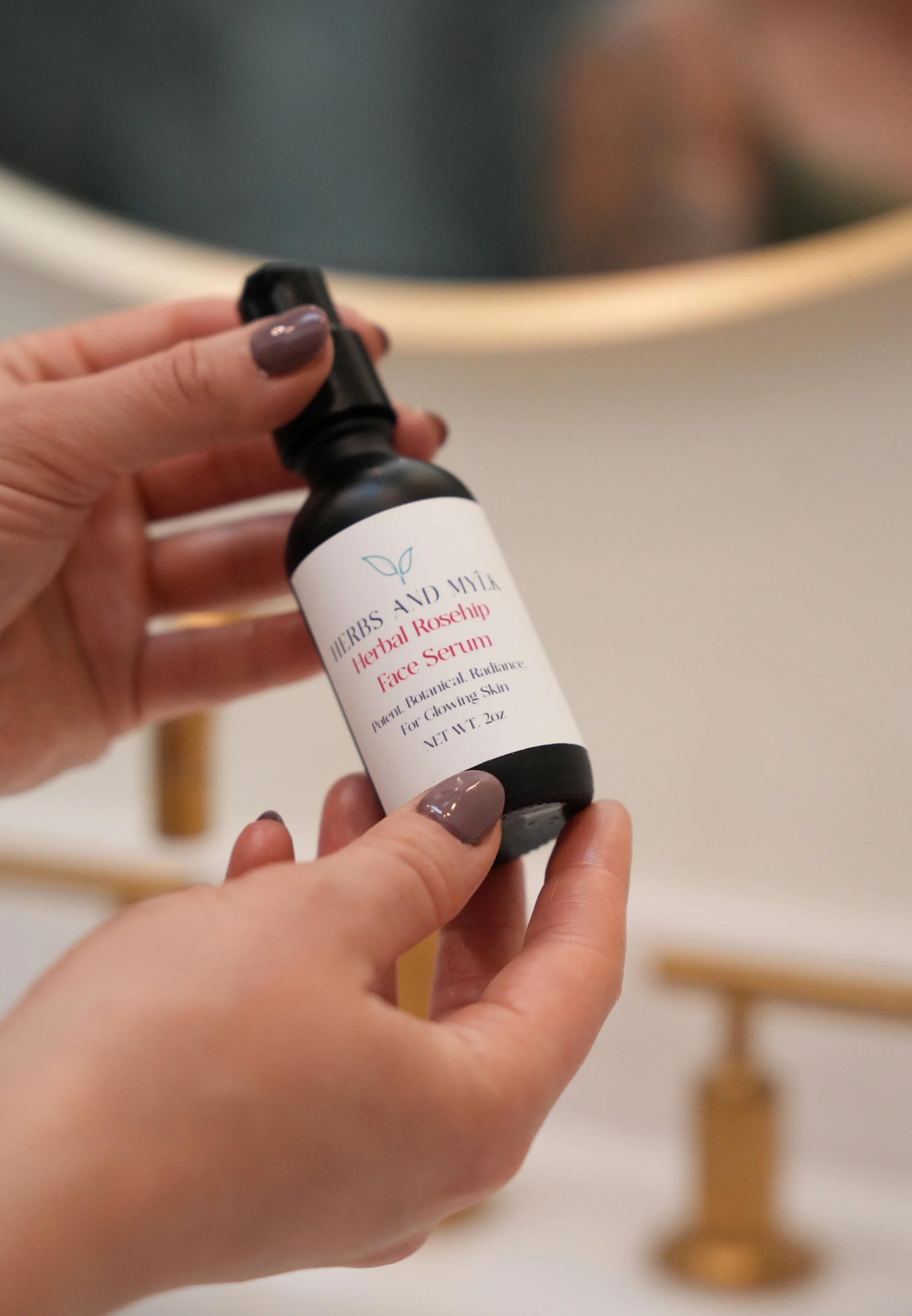 Person holding a bottle of herbs and mine herbal rosehip face serum, with a mirror and a blurred background.