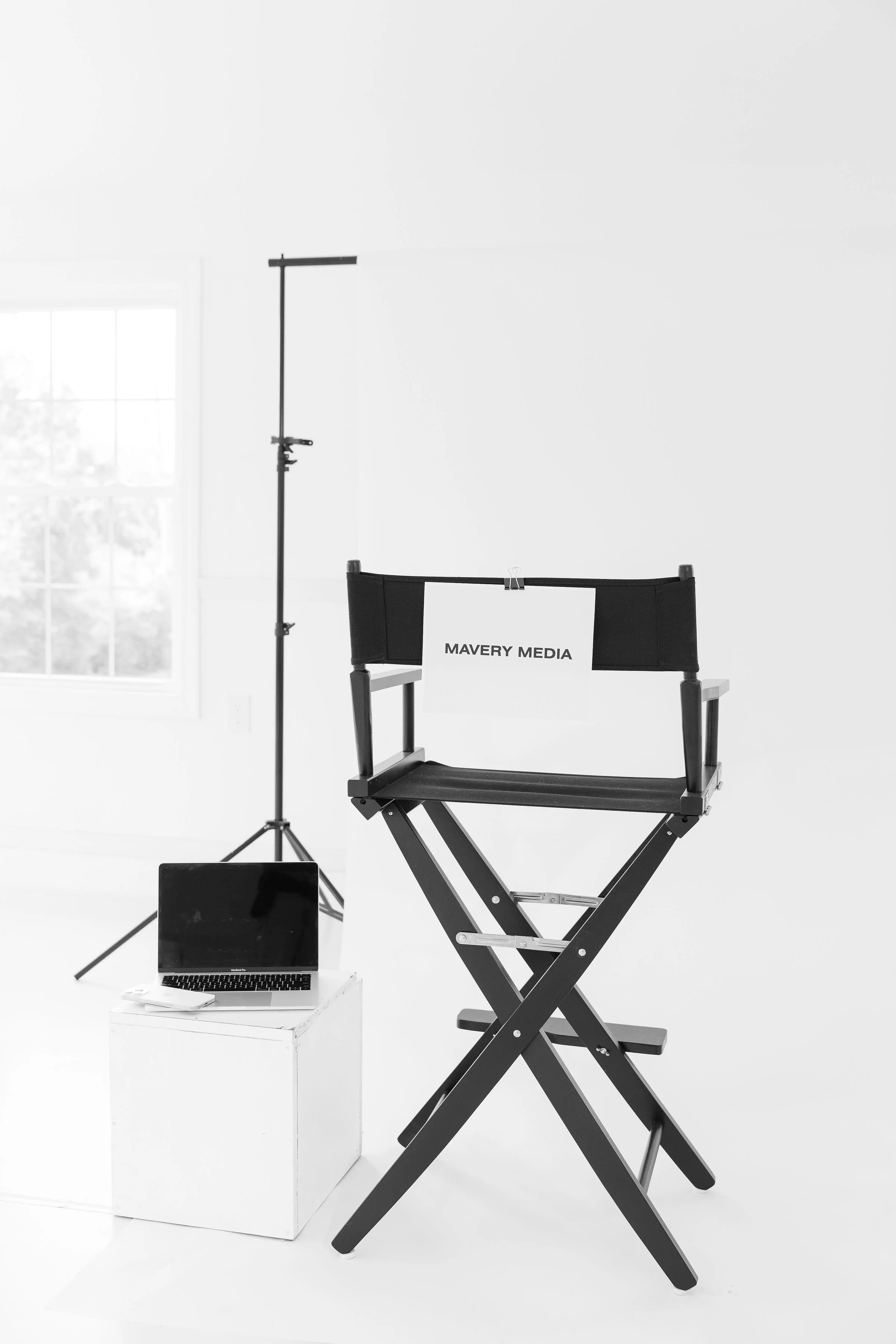 A director's chair with a sign that reads "MAVERY MEDIA," a laptop on a white stand, a smartphone, and a lighting stand in a room with a window.