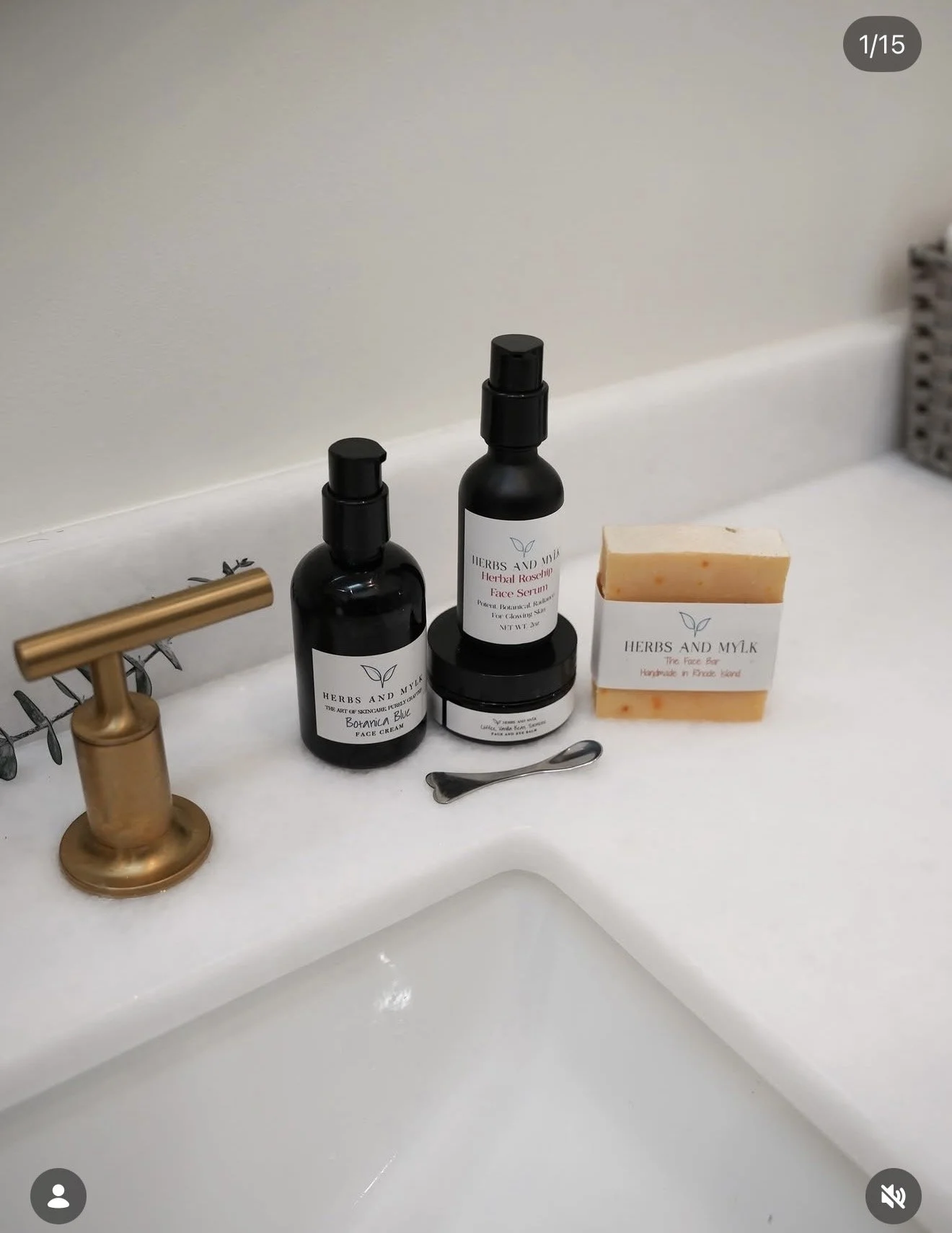 Bathroom sink countertop with a gold faucet, two black bottles labeled 'Herbs and Mylk' face serum and face cream, a bar of soap labeled 'Herbs and Mylk' herbal soap, a small metallic spoon, and a gray basket in the background.