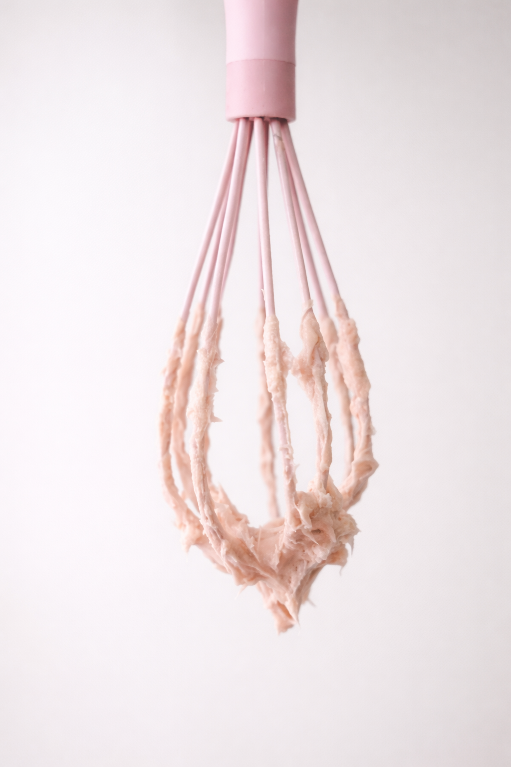 Close-up of a pink hand mixer beaters with cake batter on them, hanging against a plain white background.