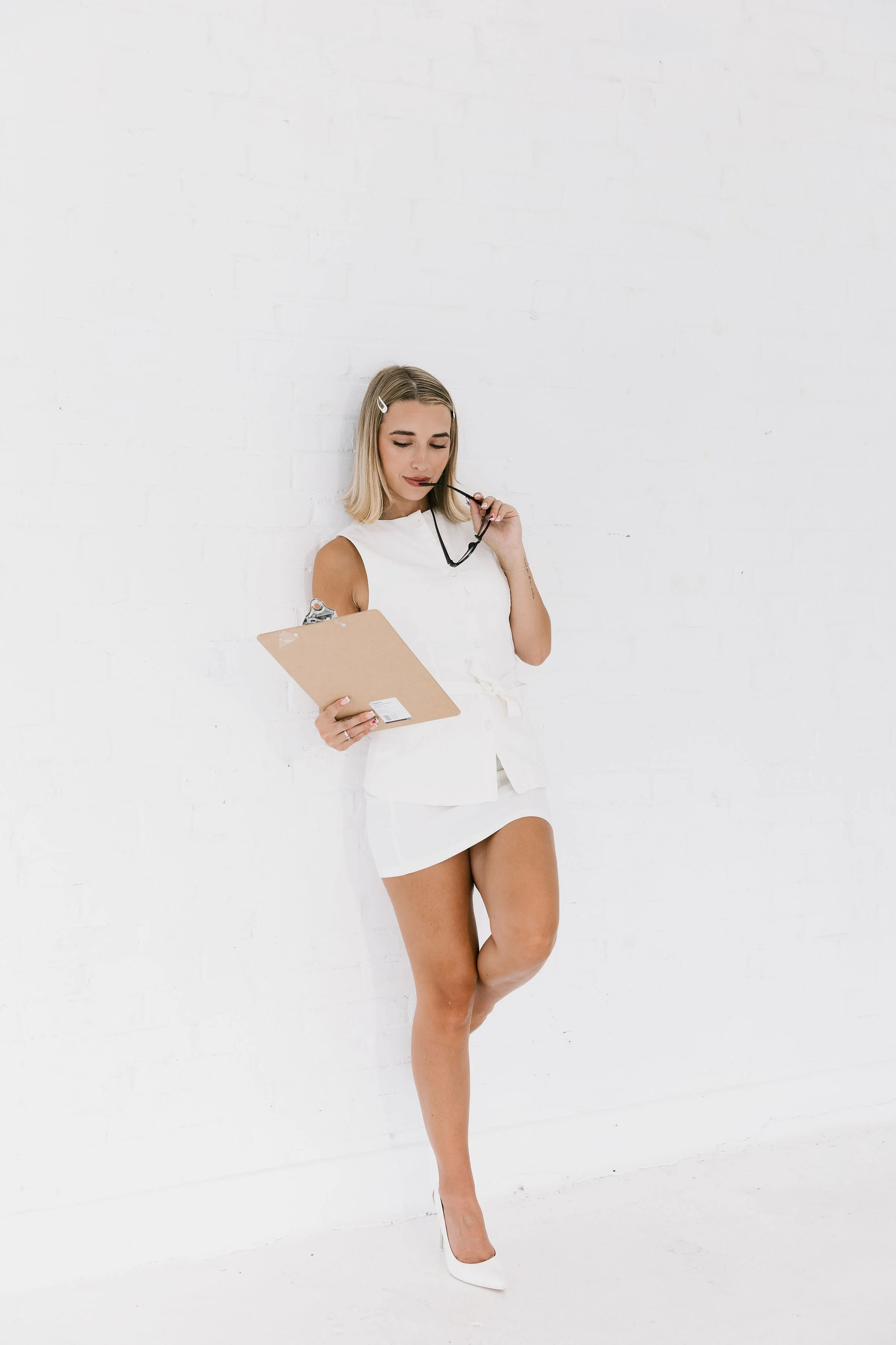 A young woman with blonde hair, wearing a sleeveless white top, white mini skirt, and white high heels, standing against a white brick wall, holding a clipboard and chewing on black-rimmed glasses.