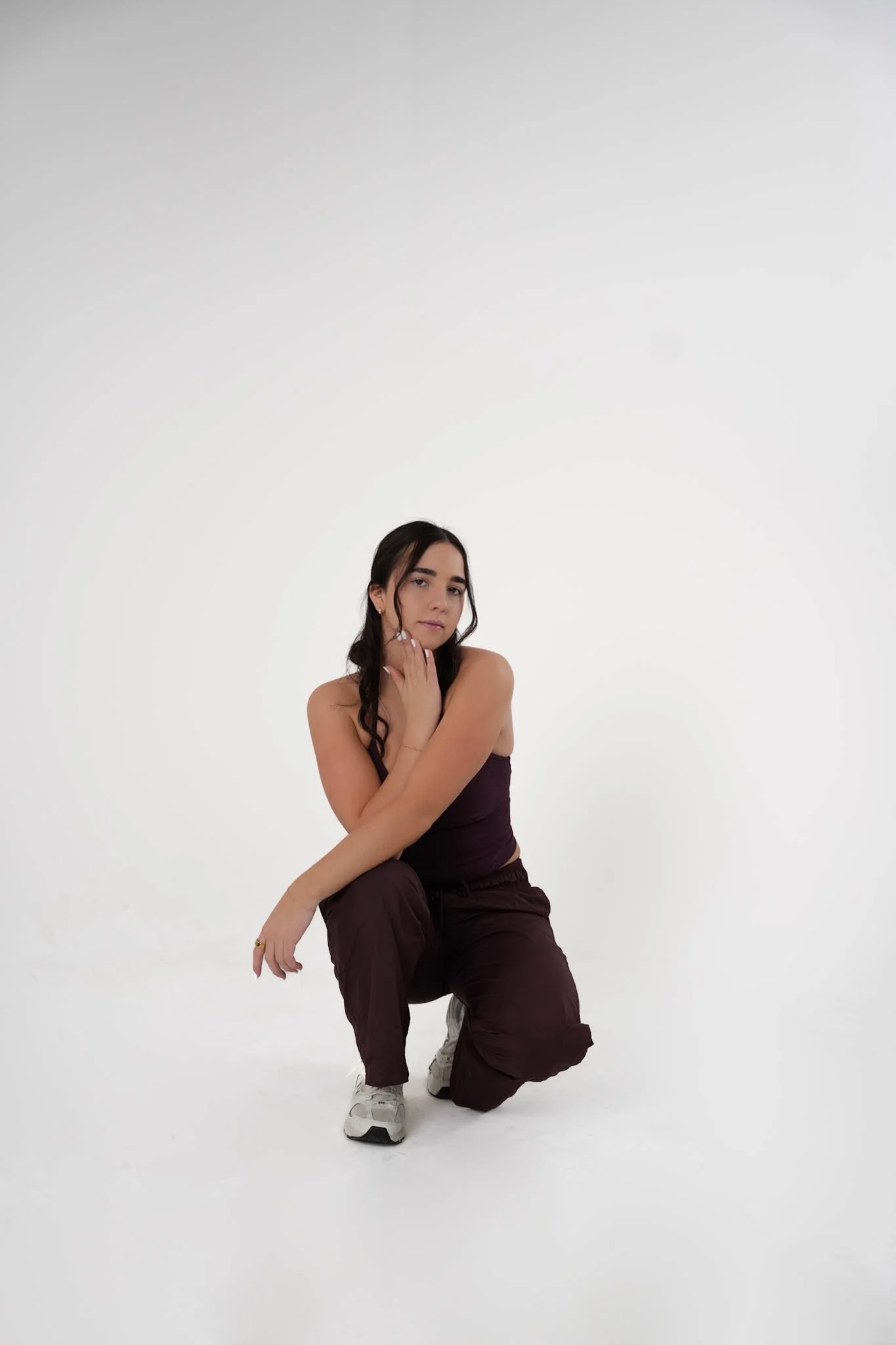 A young woman crouching against a plain white background, wearing a dark purple sleeveless top and matching pants, with long dark hair and white sneakers.