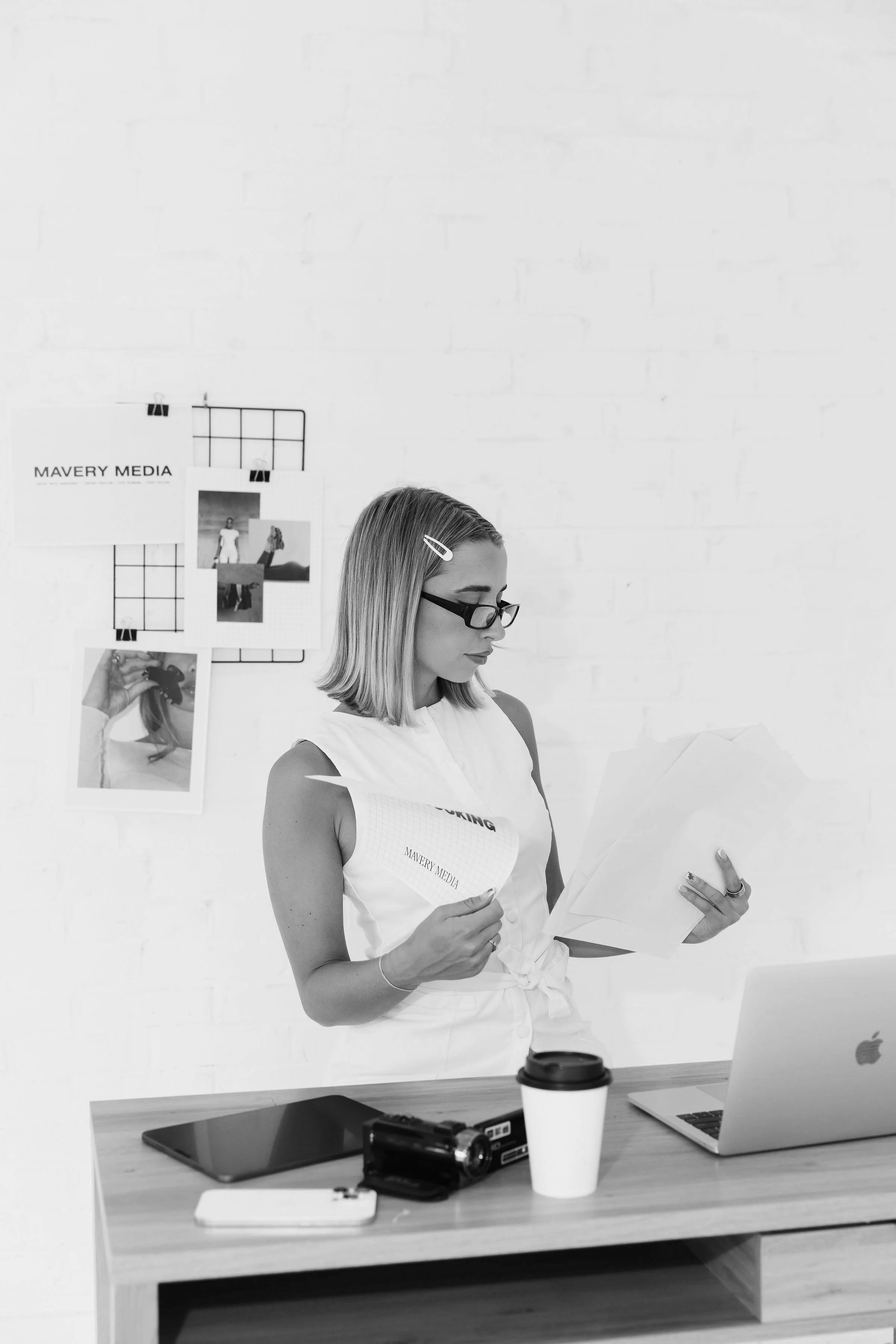 Black and white photo of a woman with glasses holding papers in an office, standing behind a desk with a laptop, coffee cup, and camera. Behind her is a wall with a grid and photos, including one labeled 'MAVER MEDIA'.