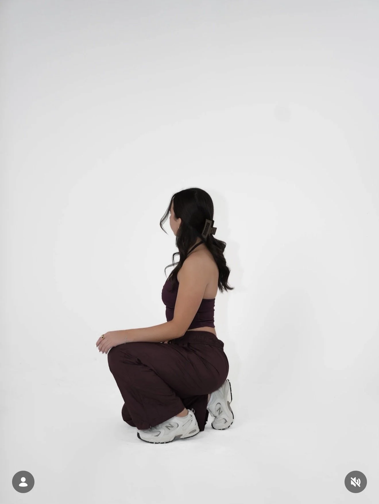 A woman with long dark hair in a high ponytail, wearing a dark purple crop top, dark purple pants, and white sneakers, is squatting in a kneeling position against a plain white background.