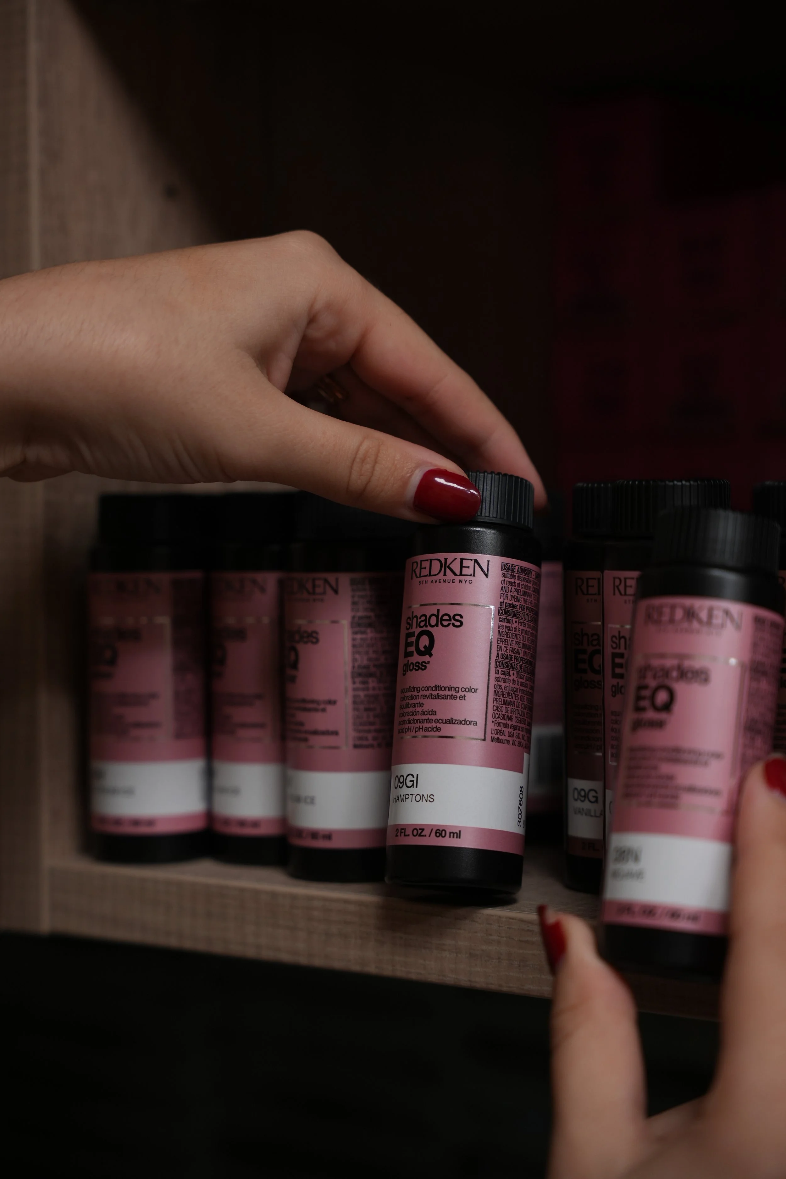 A person's hand with painted red nails reaching for a bottle of Redken shades EQ gloss hair color among other similar bottles on a wooden shelf.