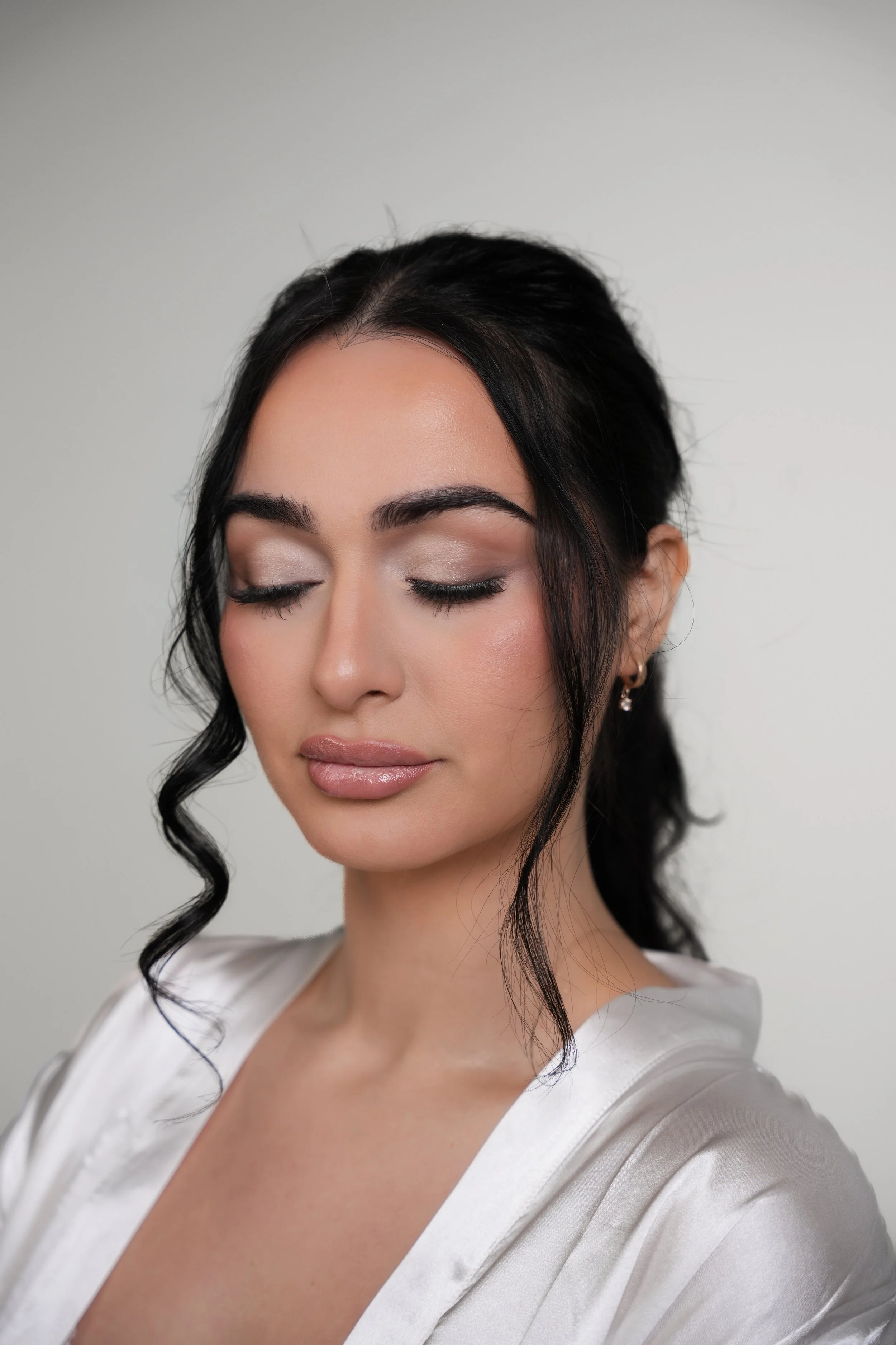 A woman with closed eyes, dark wavy hair, and light makeup, wearing a white satin robe, against a plain light gray background.