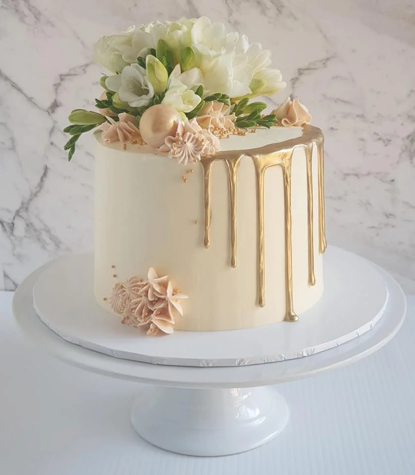Elegant white cake decorated with white flowers, peach-colored piped buttercream, and gold drip glaze on a white cake stand against a marble background.
