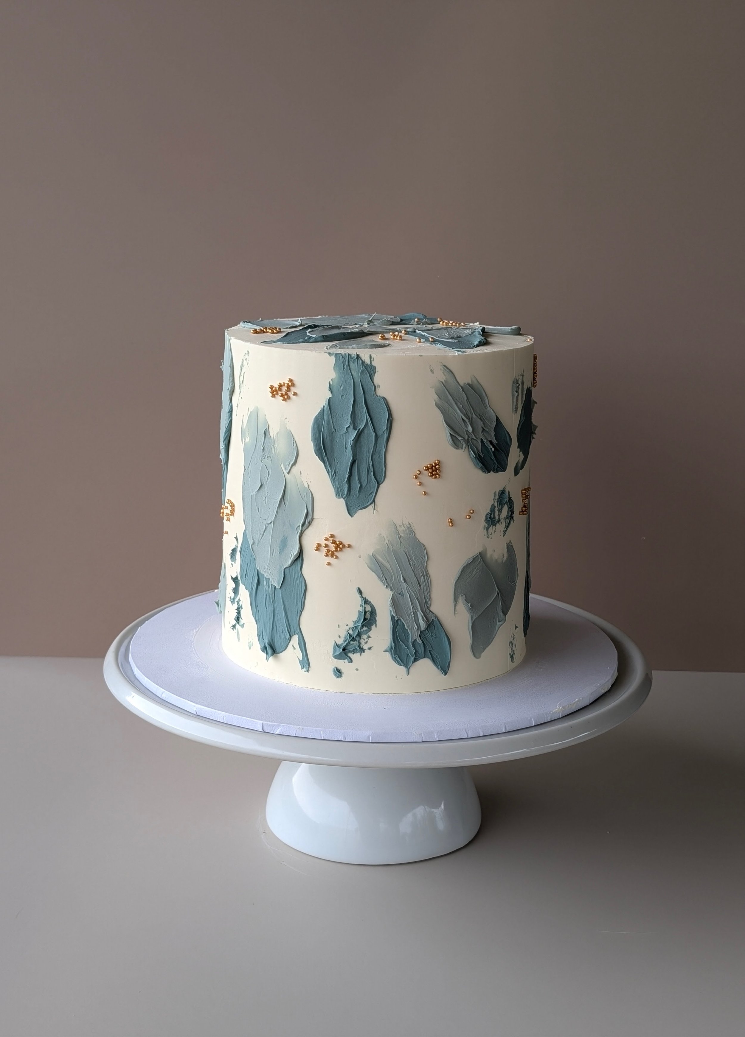 A white cake with blue and gray textured buttercream decorations and small gold beads, displayed on a white cake stand against a neutral background.