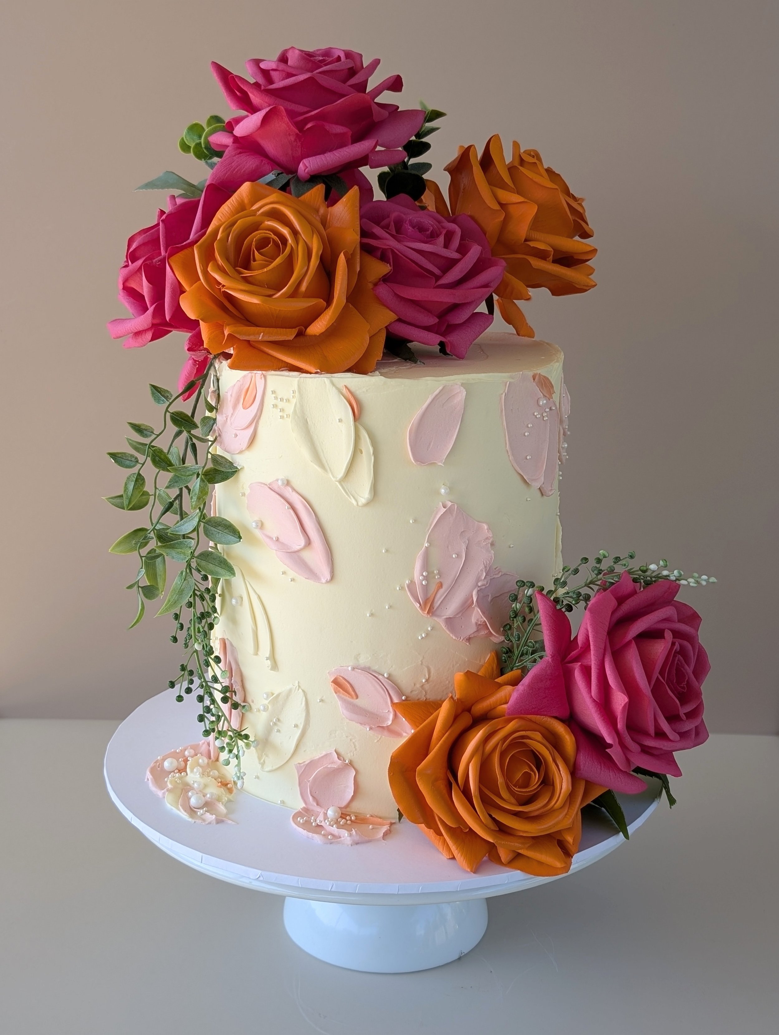 A tall, elegant wedding cake decorated with pink and orange roses, green leaves, and pink petal-like frosting accents on a white cake stand.