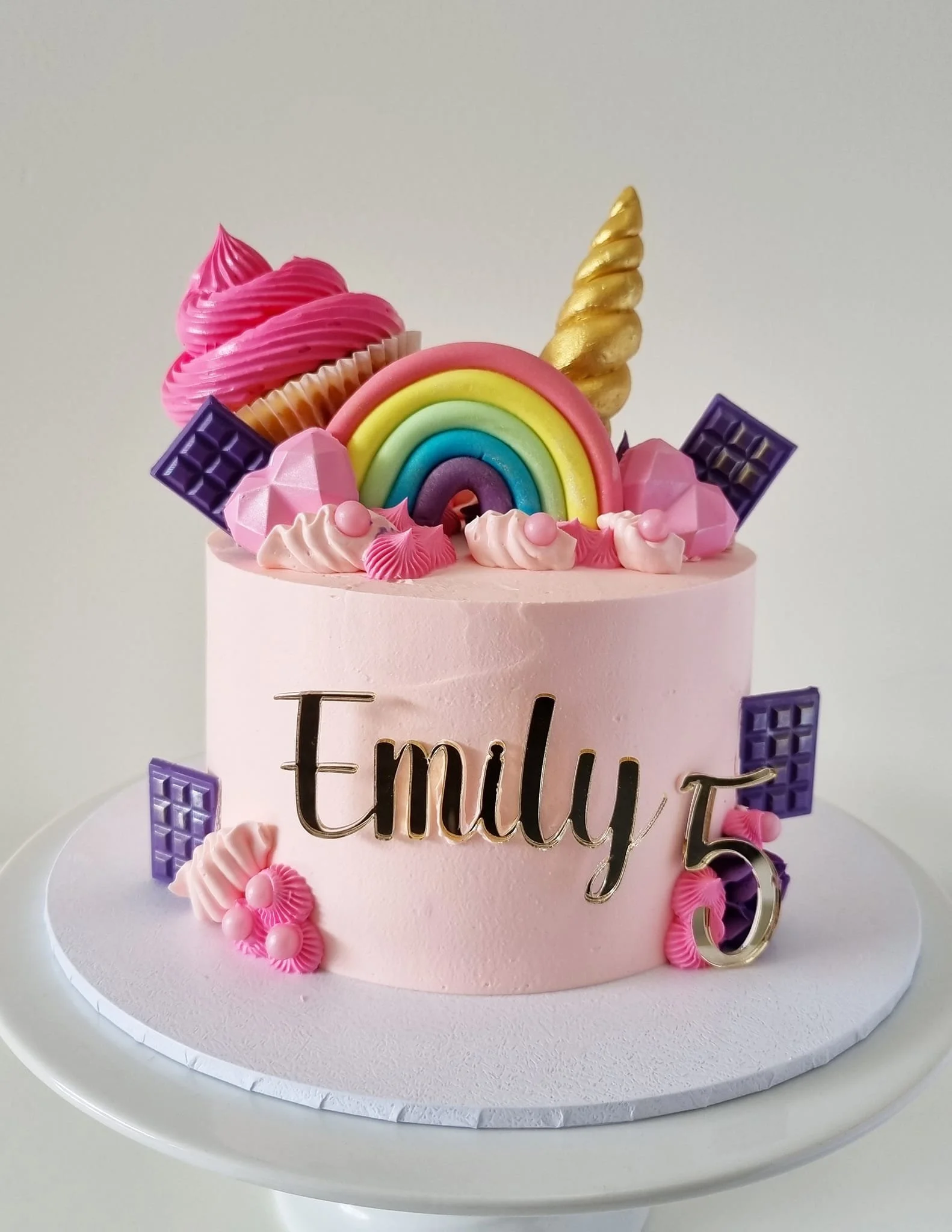 Pink birthday cake with colorful rainbow, unicorn horn, and decorative chocolates, personalized with the name Emily and the number 5.