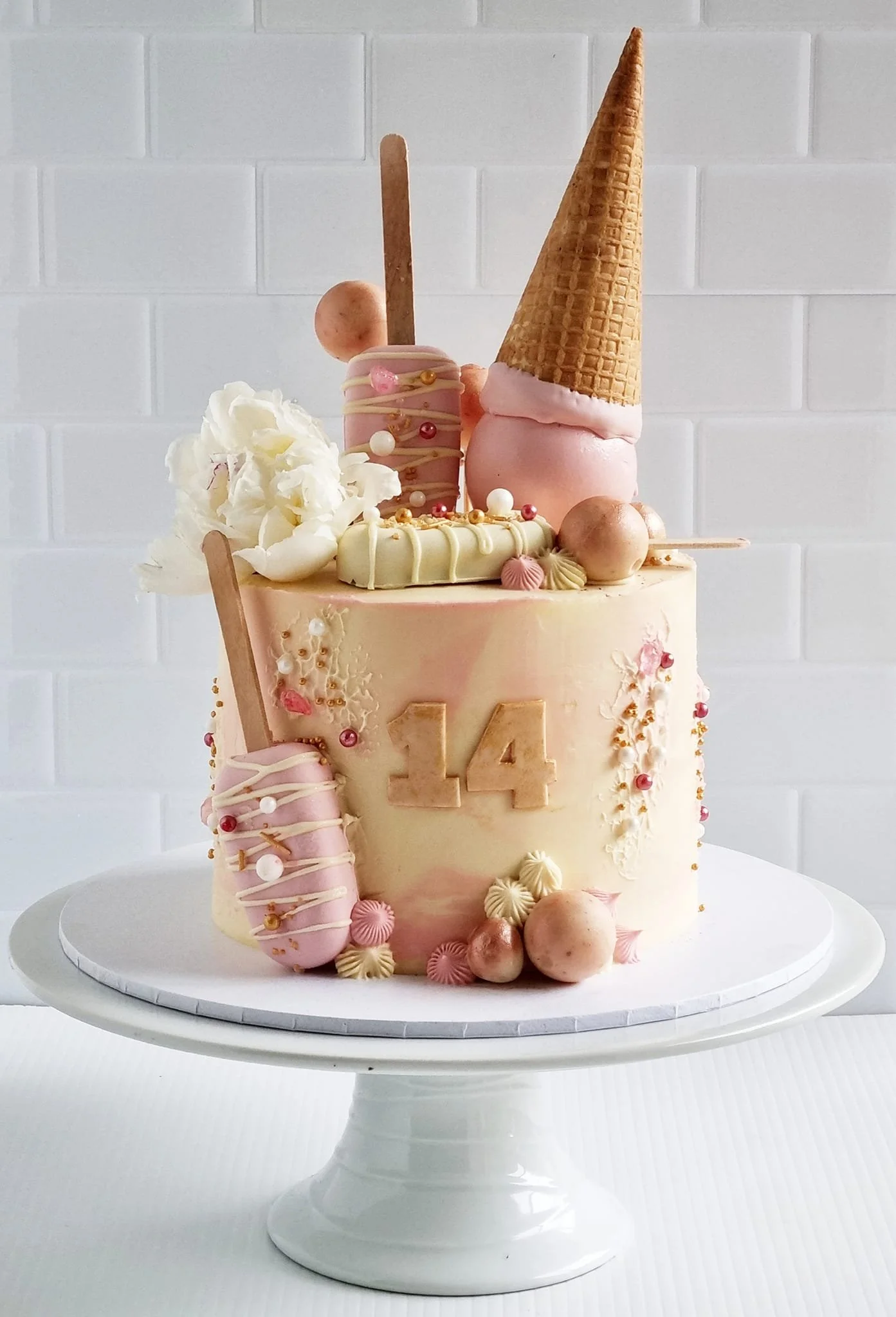 A pink and white decorated birthday cake with the number 14 on it, topped with ice cream cones, macarons, and various candies.