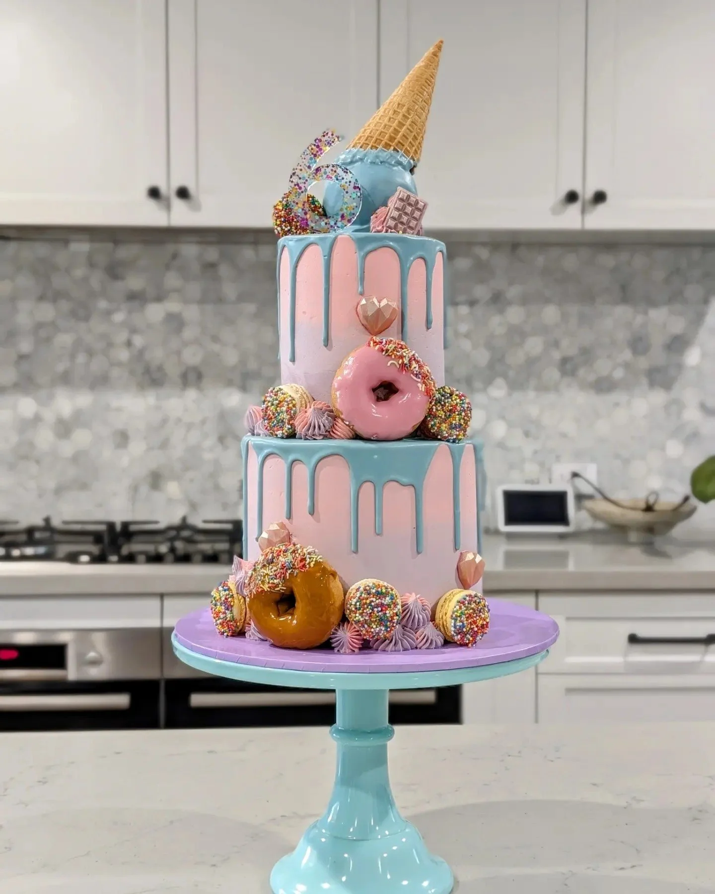 Three-tiered pastel pink and blue drip cake decorated with donuts, sprinkles, a waffle cone, and various colorful candies, placed on a light blue cake stand in a modern kitchen.