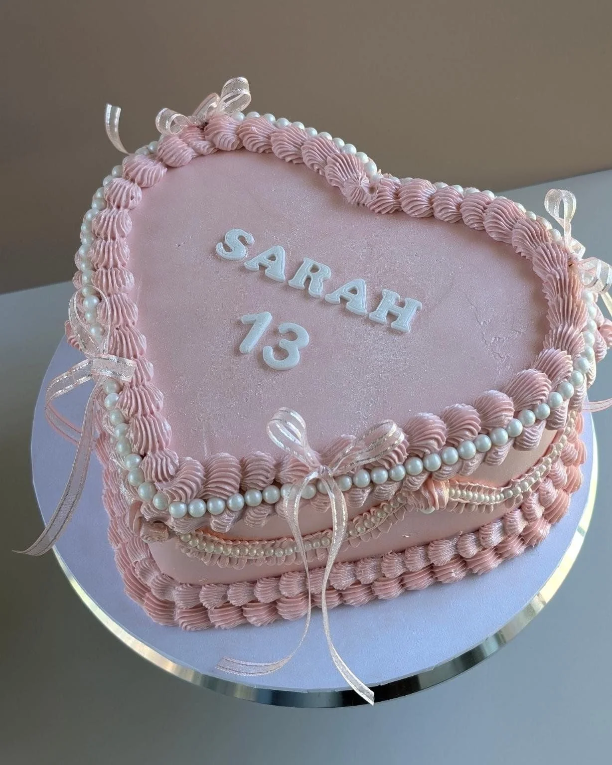 A pink heart-shaped birthday cake decorated with piped pink frosting, white pearls, and ribbons. It has the name 'Sarah' and the age '13' written on top in white icing.