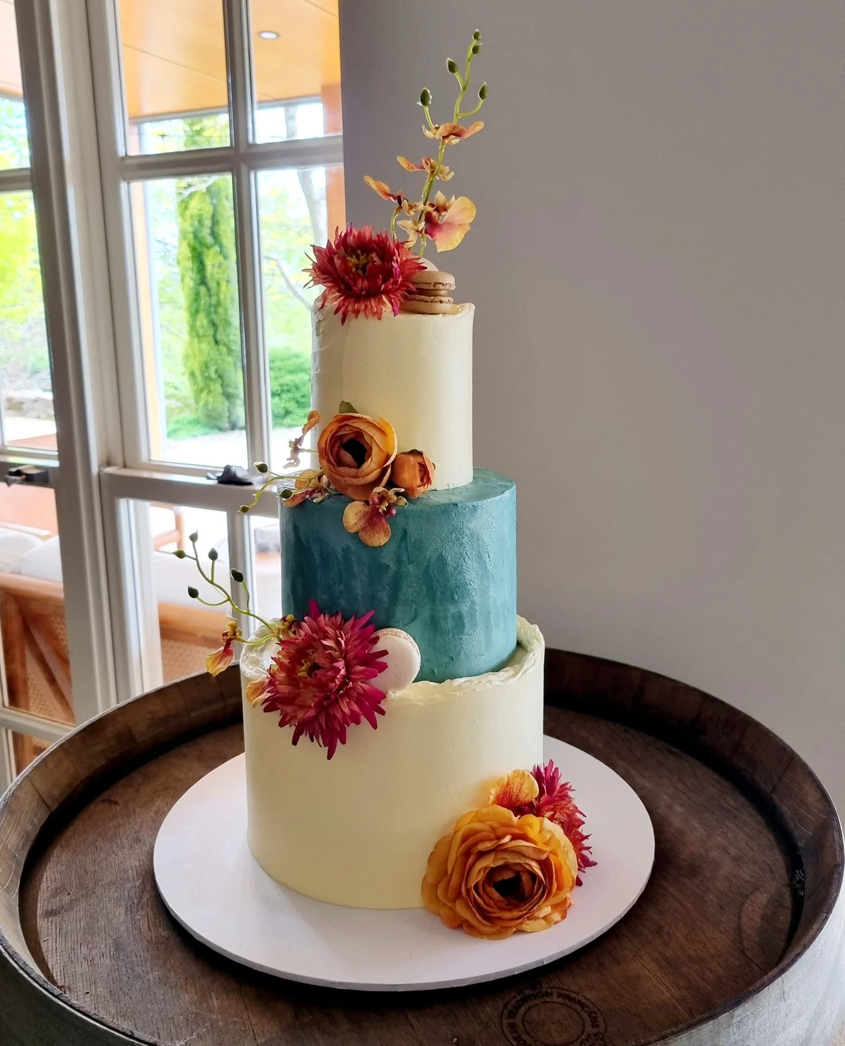 Three-tiered wedding cake with colorful flowers, placed on a wooden tray near a window.