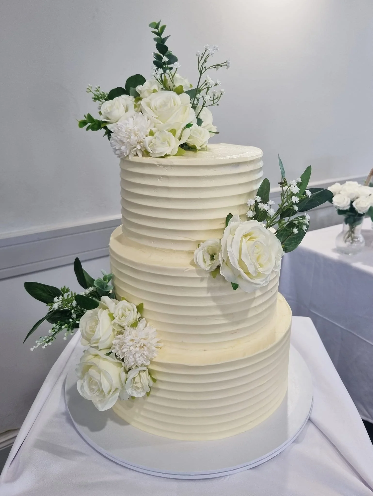 Three-tier white wedding cake decorated with white roses, carnations, and greenery.