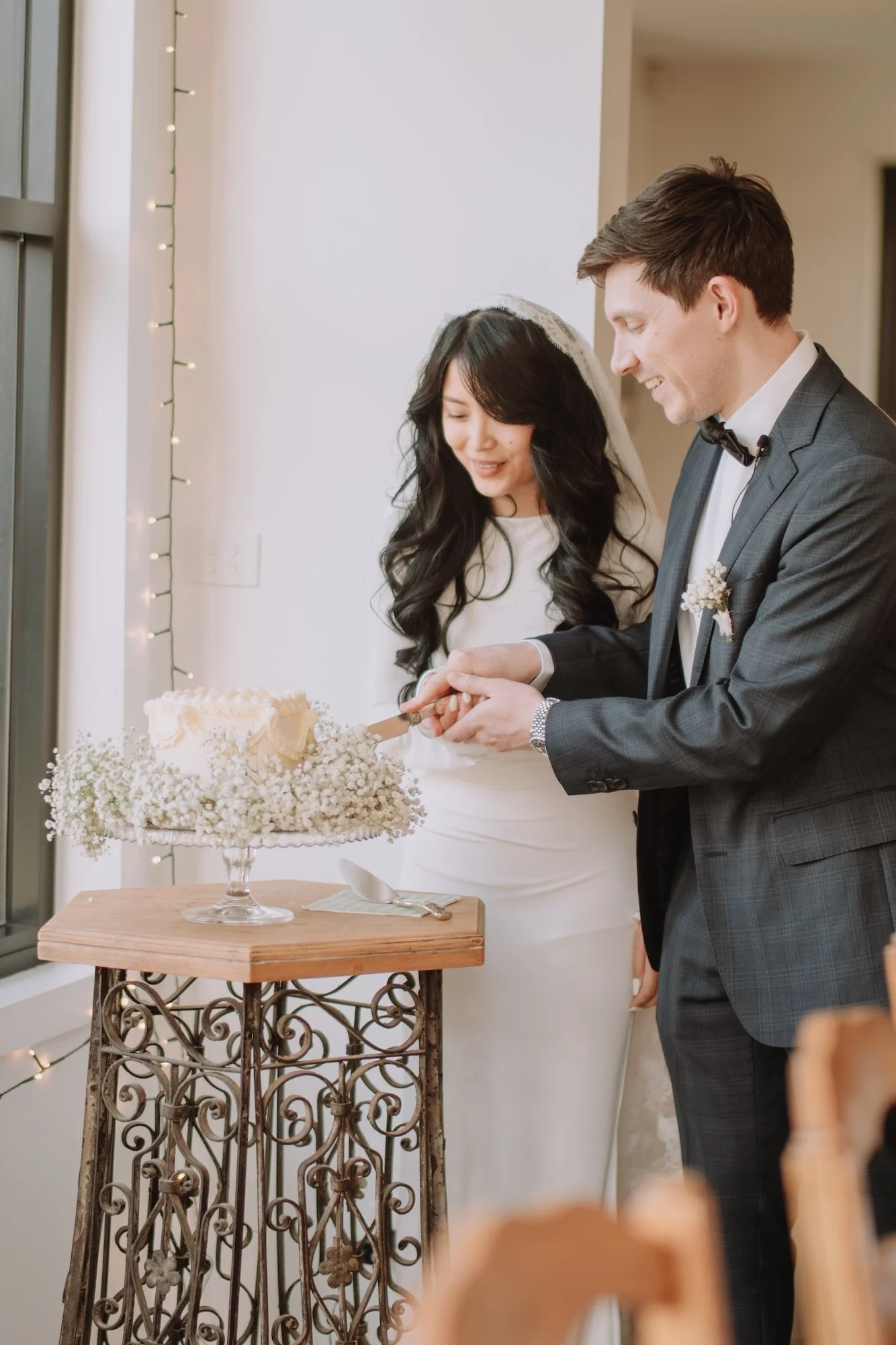 A newlywed couple, a woman with long black hair in a white wedding dress and a man in a gray suit, cutting a wedding cake together during their wedding reception.