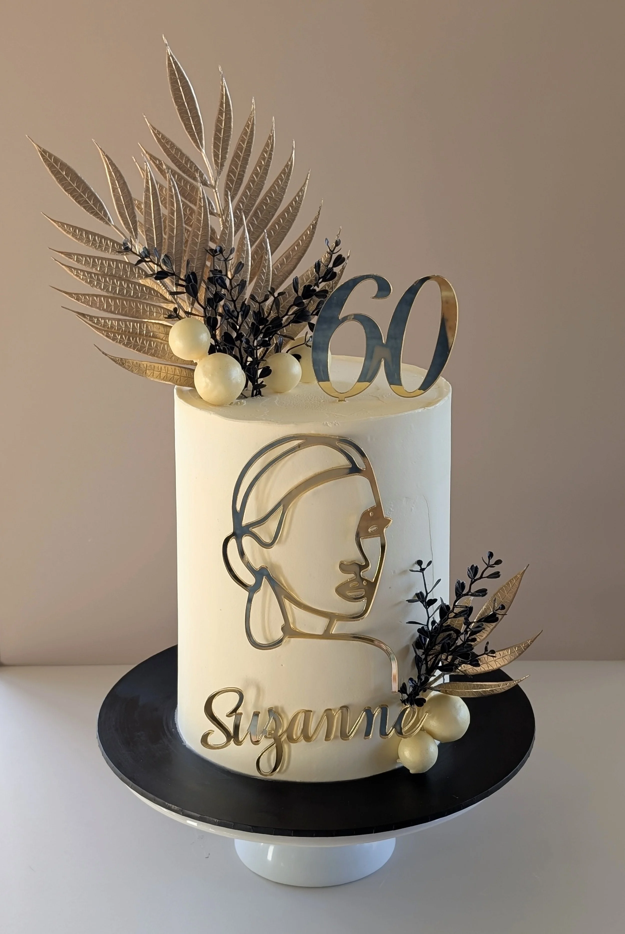 Elegant white birthday cake with gold and black decorations, floral accents, a gold outline of a woman's face, the number 60, and the name Suzanne on it.