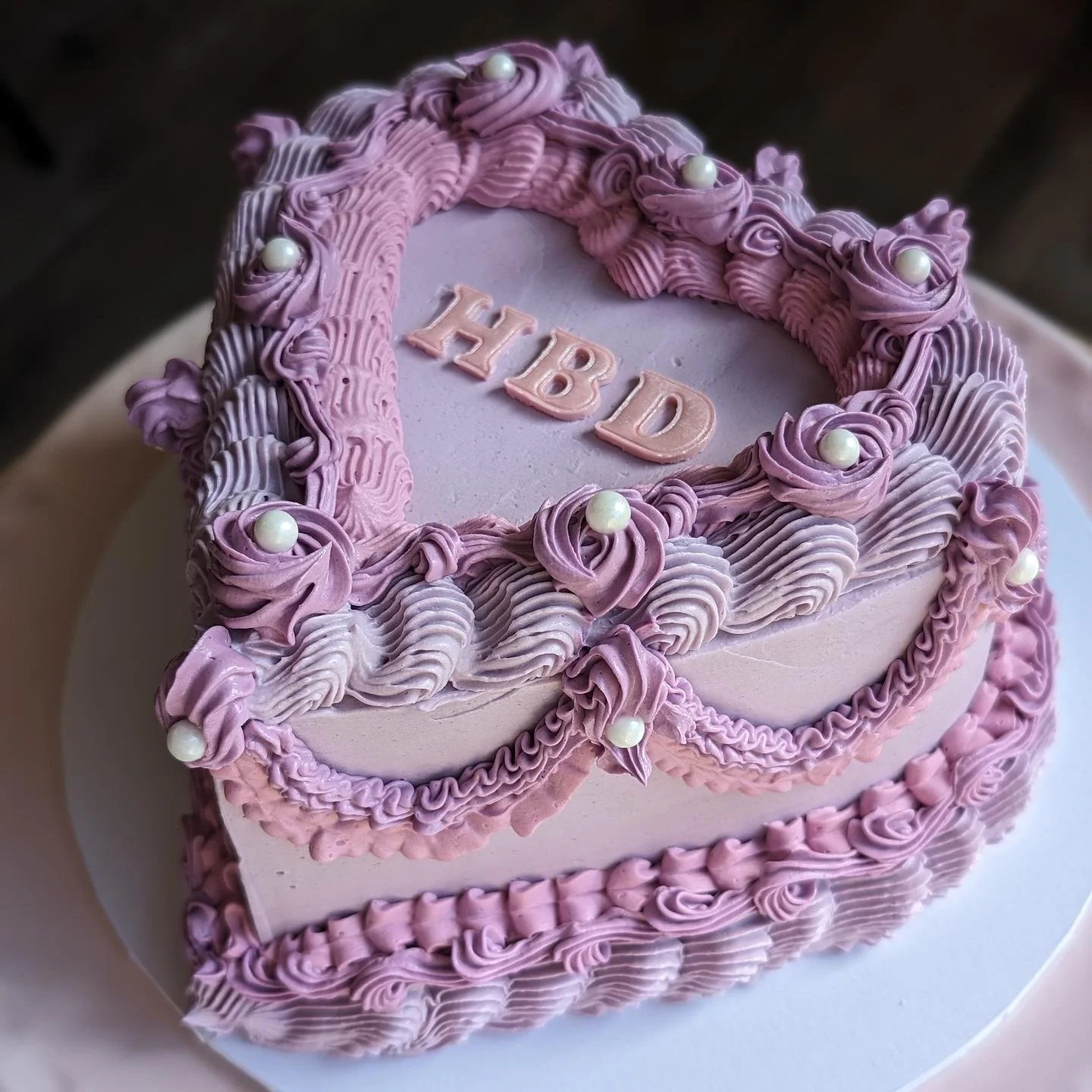 A heart-shaped birthday cake decorated with pink and purple frosting and white pearls, with the word "HBD" written on top.