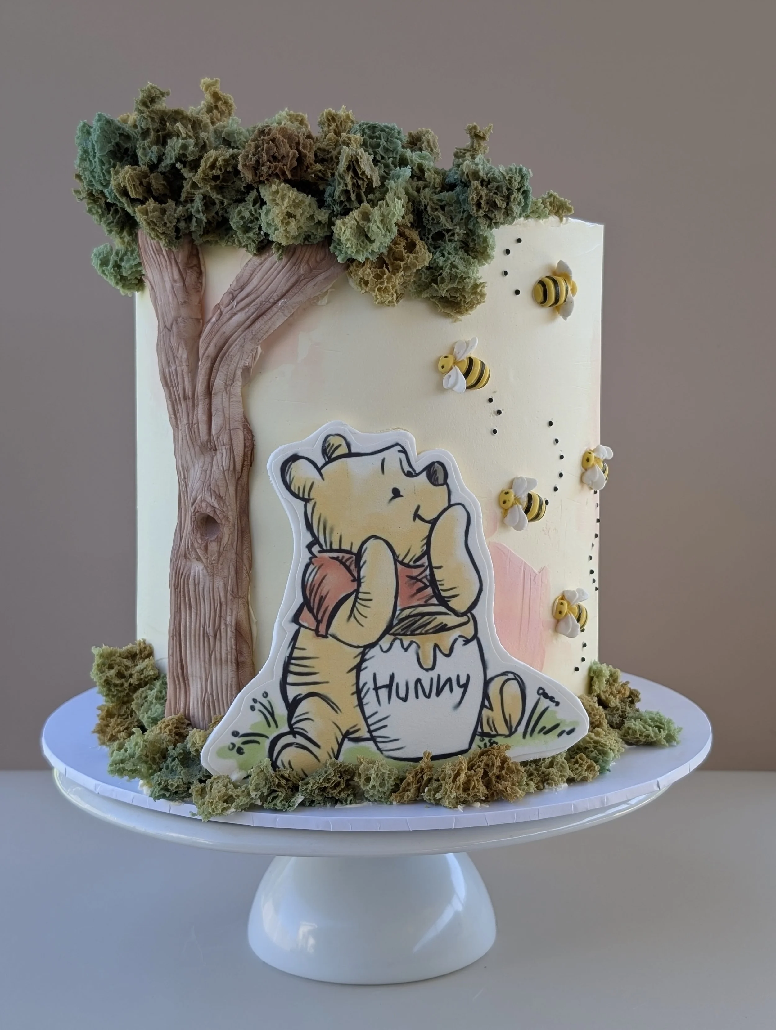 Cake decorated with a Winnie the Pooh theme, featuring a tree made of textured green and brown material, bee decorations, and a fondant figure of Winnie the Pooh holding a honey pot labeled 'Hunny'.