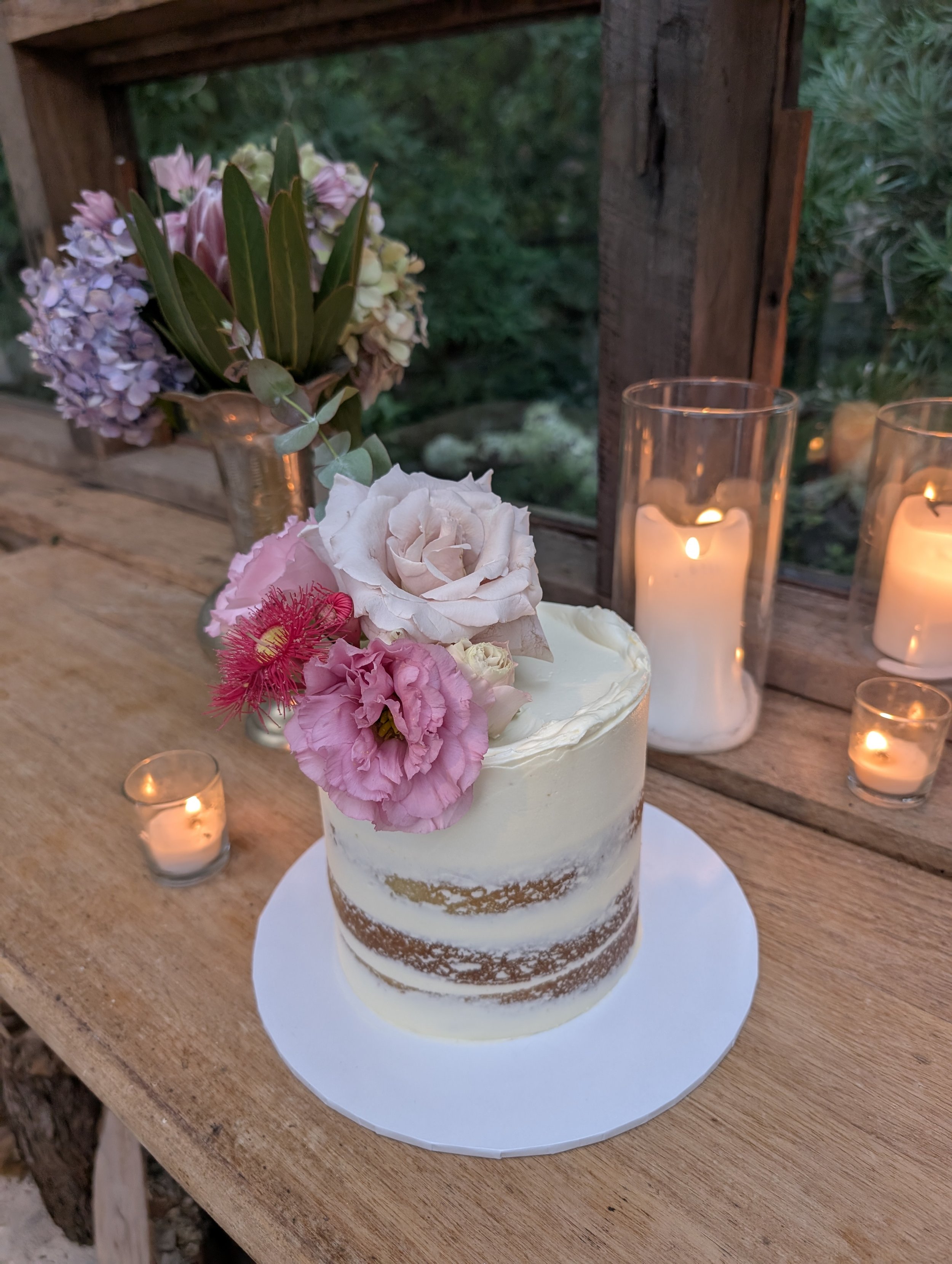 A semi-naked cake decorated with pink and white roses and lisianthus flowers, set on a rustic wooden table with candles and a flower arrangement in the background.