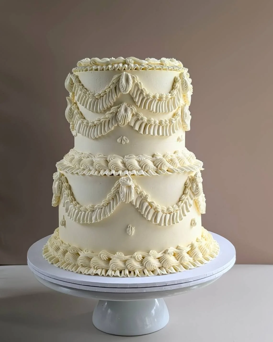 A three-tier white vintage Lambeth wedding cake decorated with piped cream swags and small cream rosettes, displayed on a white cake stand.