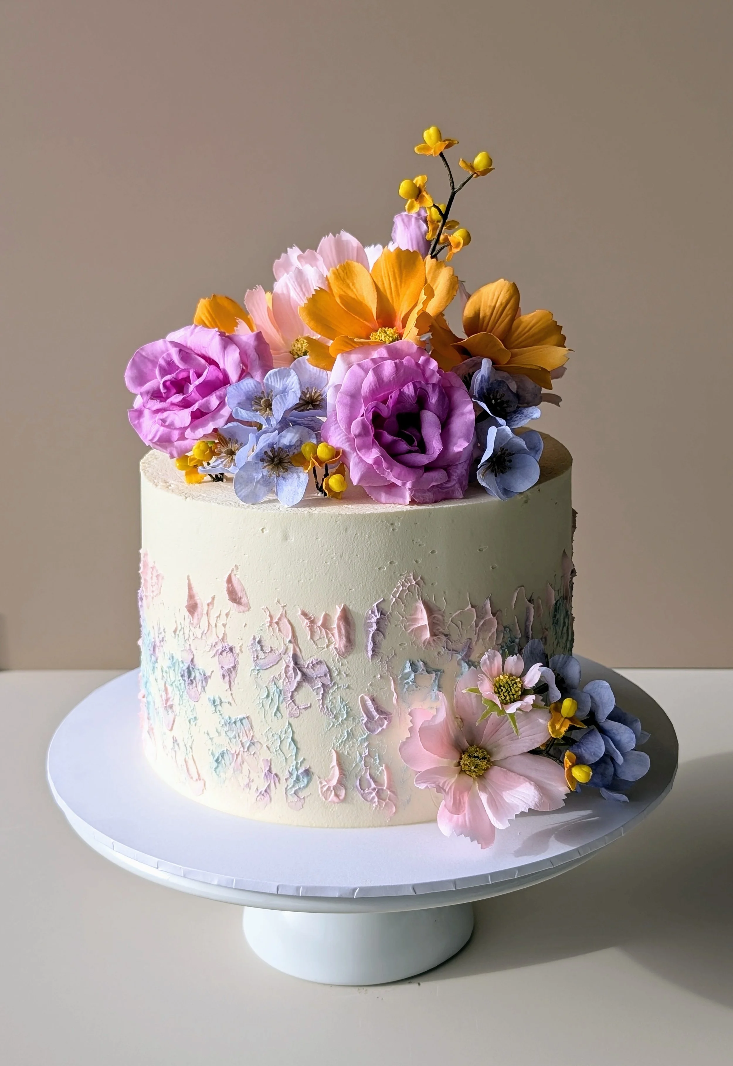A round cake with textured, pastel-colored icing decorated with pink, purple, yellow, and blue flowers on top and on the side, placed on a white cake stand.