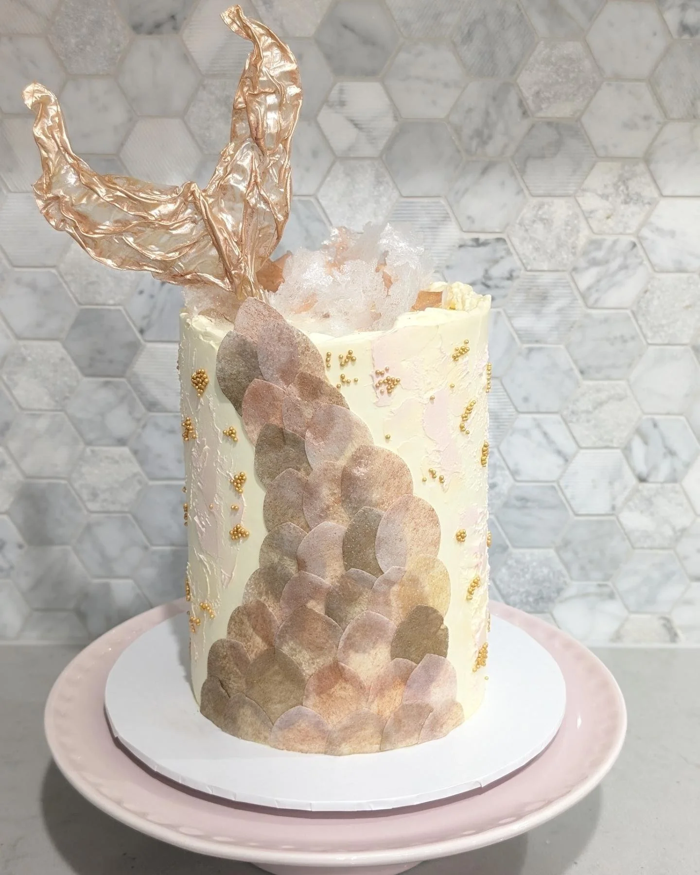 A tall, cylindrical cake decorated with gold and pastel-colored edible accents, topped with a metallic gold leaf decoration. The cake features a pattern of overlapping circles in soft hues on its side, with small gold edible beads scattered on the su