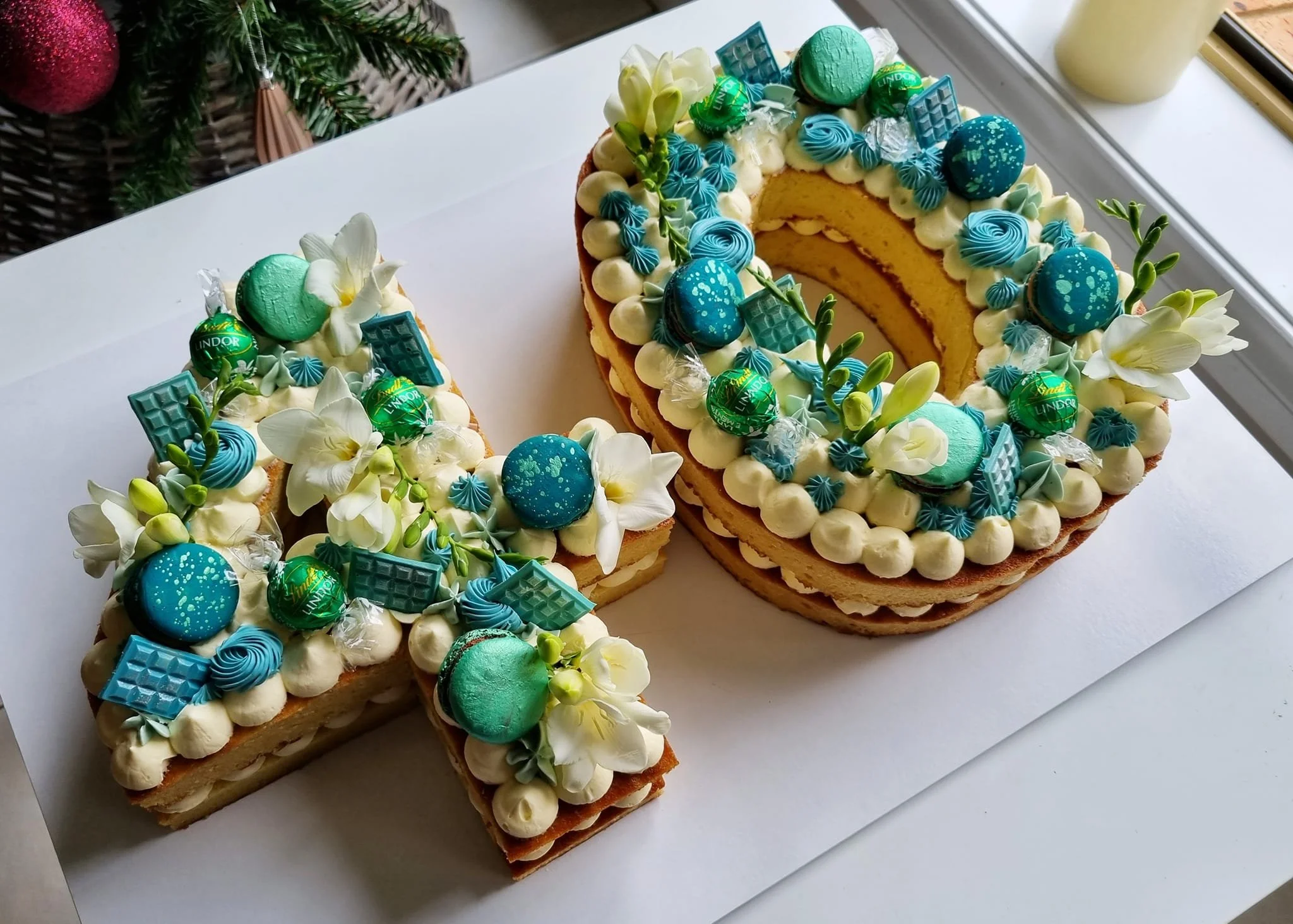 A decorated cake shaped as the number 4, with white flowers, blue decorative elements, and green candies, placed on a white surface near a window.