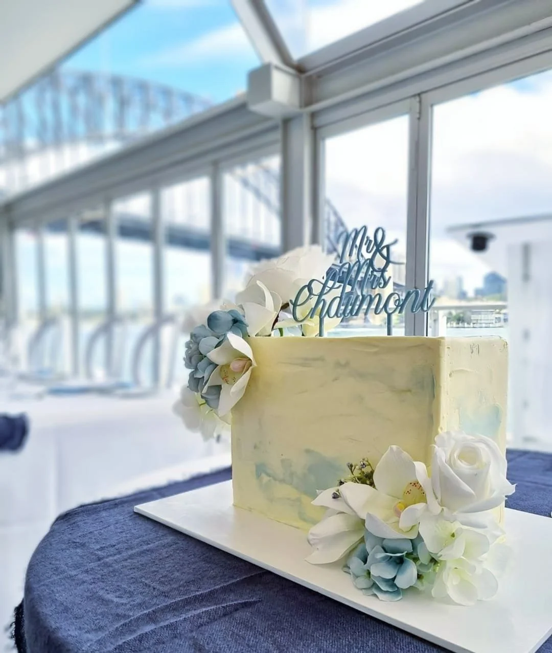 Wedding cake with white and light blue flowers, and a 'Mrs. and Mr. Chaimont' cake topper, on a dark table near large windows with Sydney Harbour Bridge cityscape views.