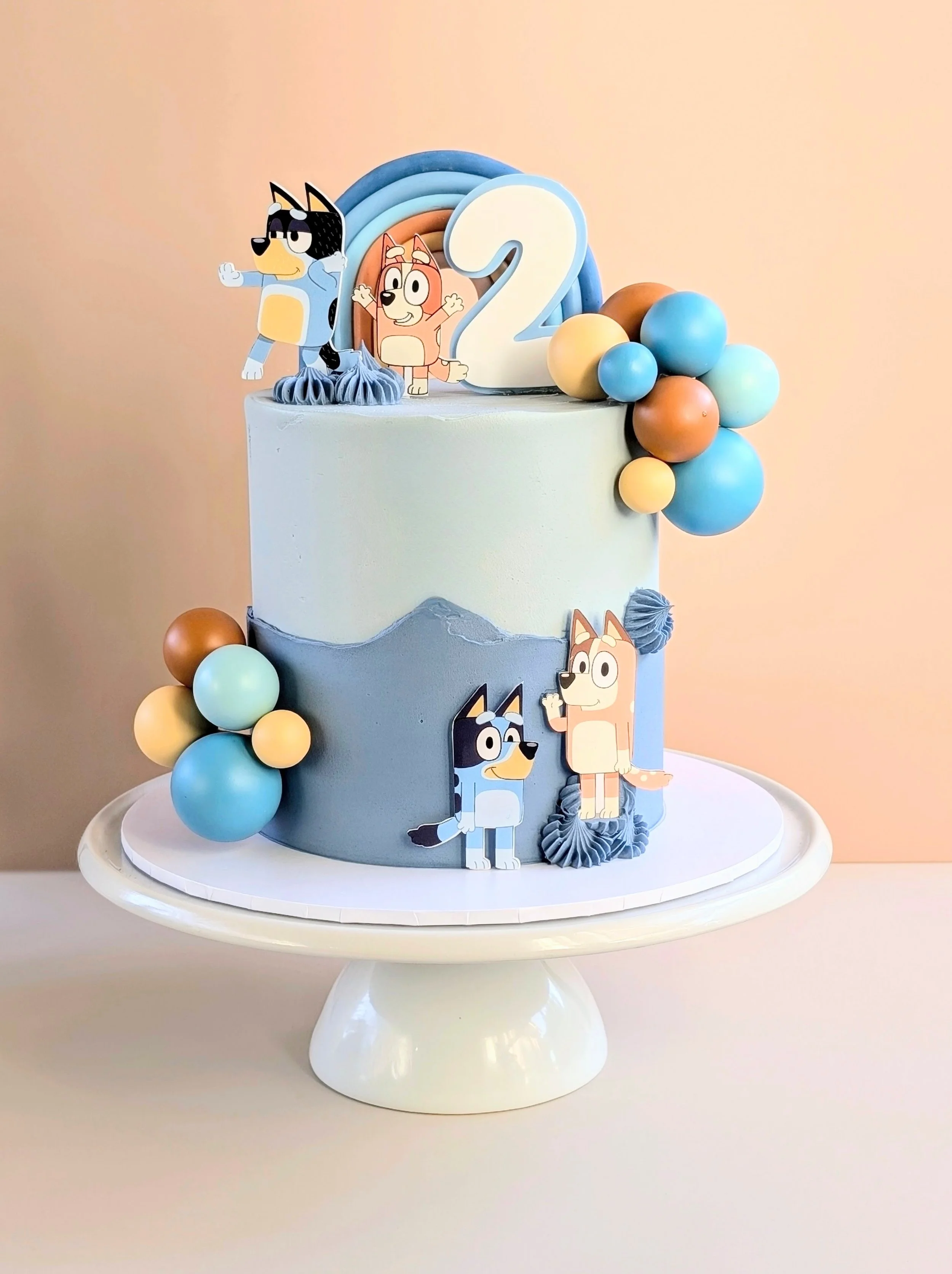 Two-tiered birthday cake decorated with cartoon dog characters, colorful balloons, and a large number 2 topper, set on a white cake stand.