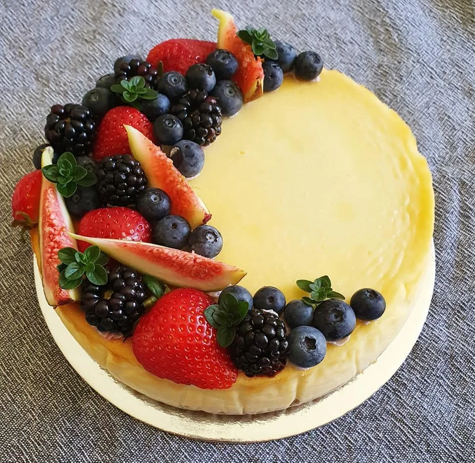 A round cake decorated with fresh strawberries, blueberries, blackberries, sliced figs, and small green leaves, with a smooth yellow frosting.
