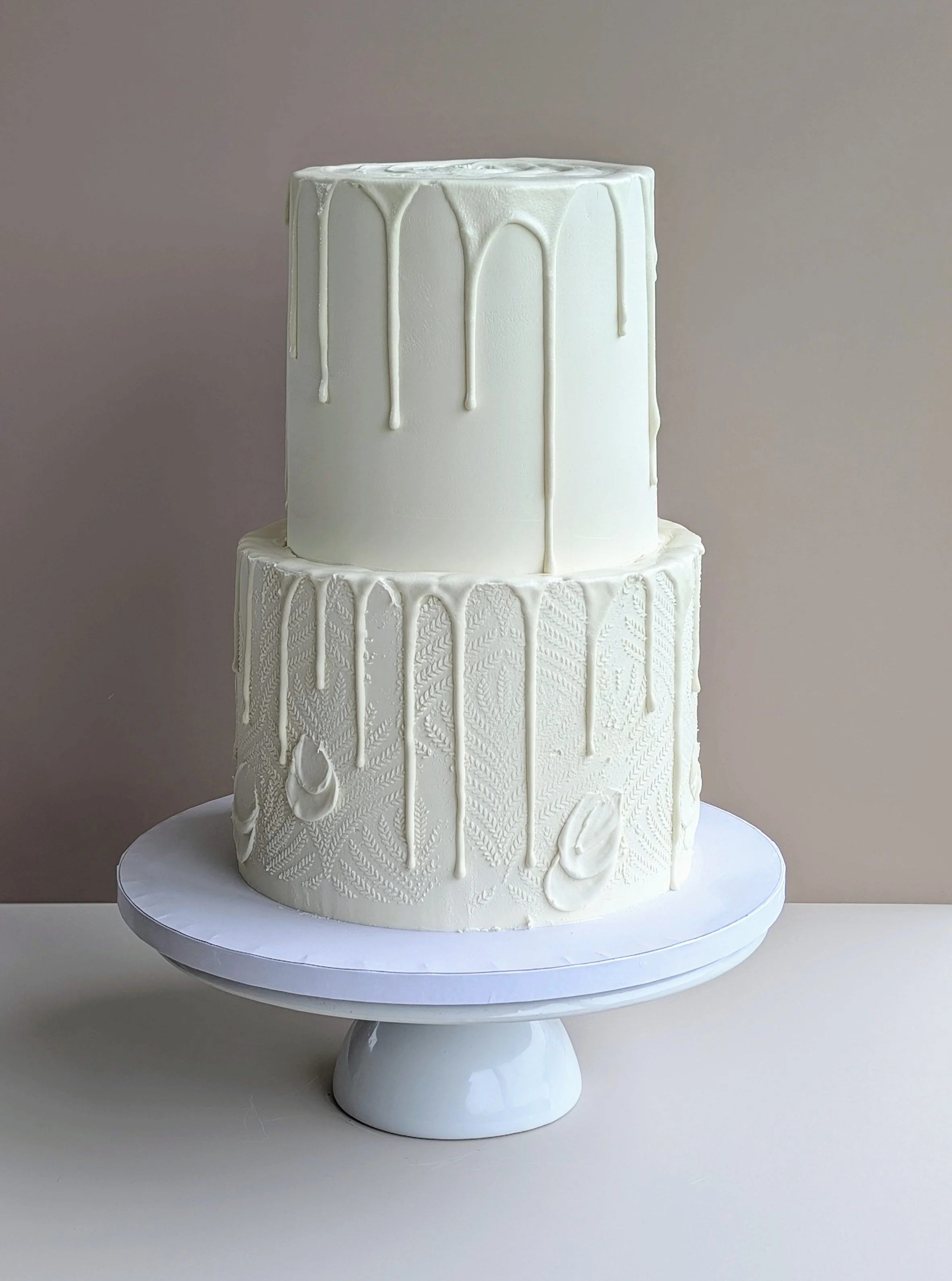Two-tiered white cake with a subtle lace pattern and white drip decorations on a white cake stand against a neutral background.