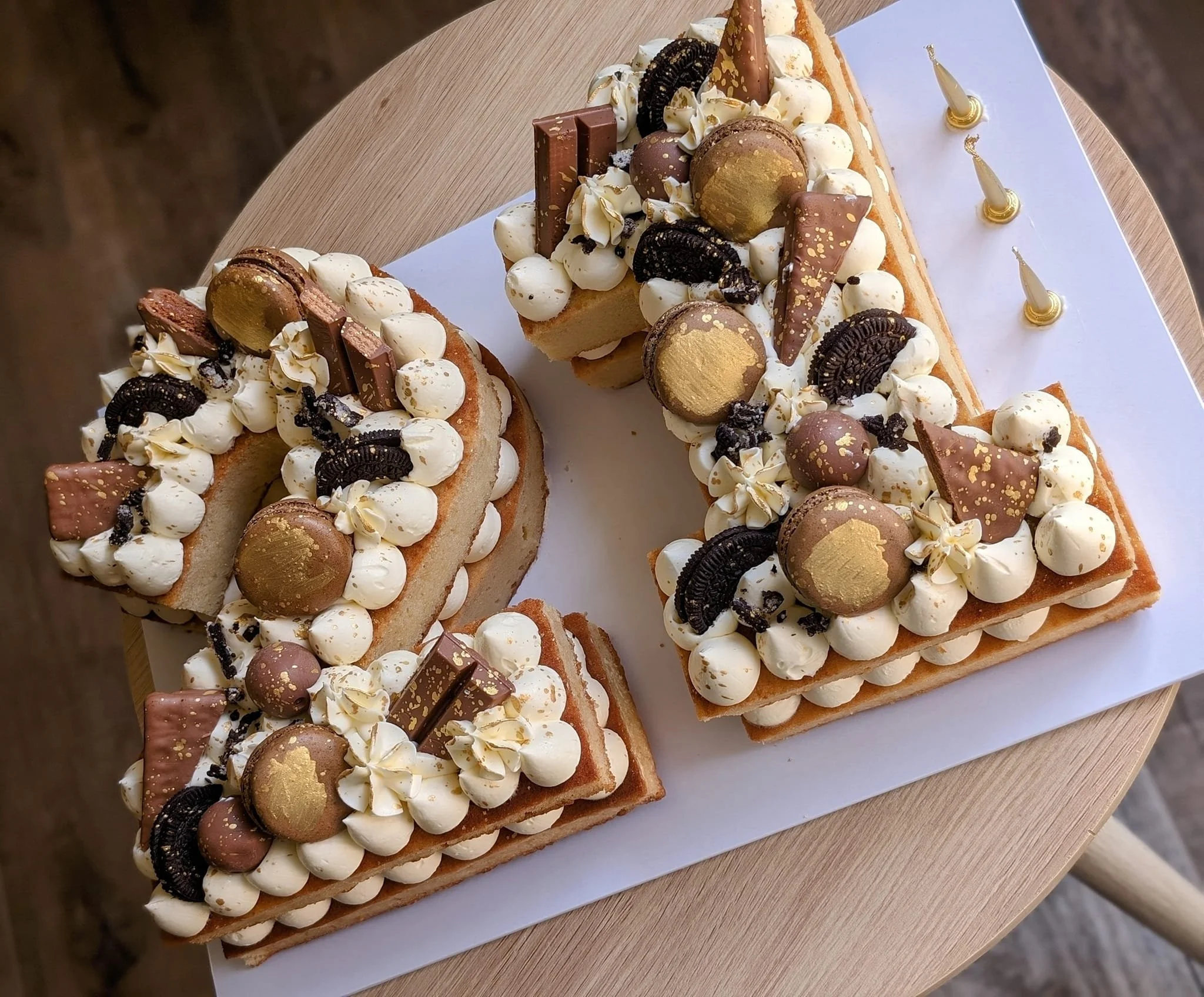 A decorative cake decorated with white frosting, chocolate pieces, cookies, and gold accents, arranged on a white board on a round wooden table.