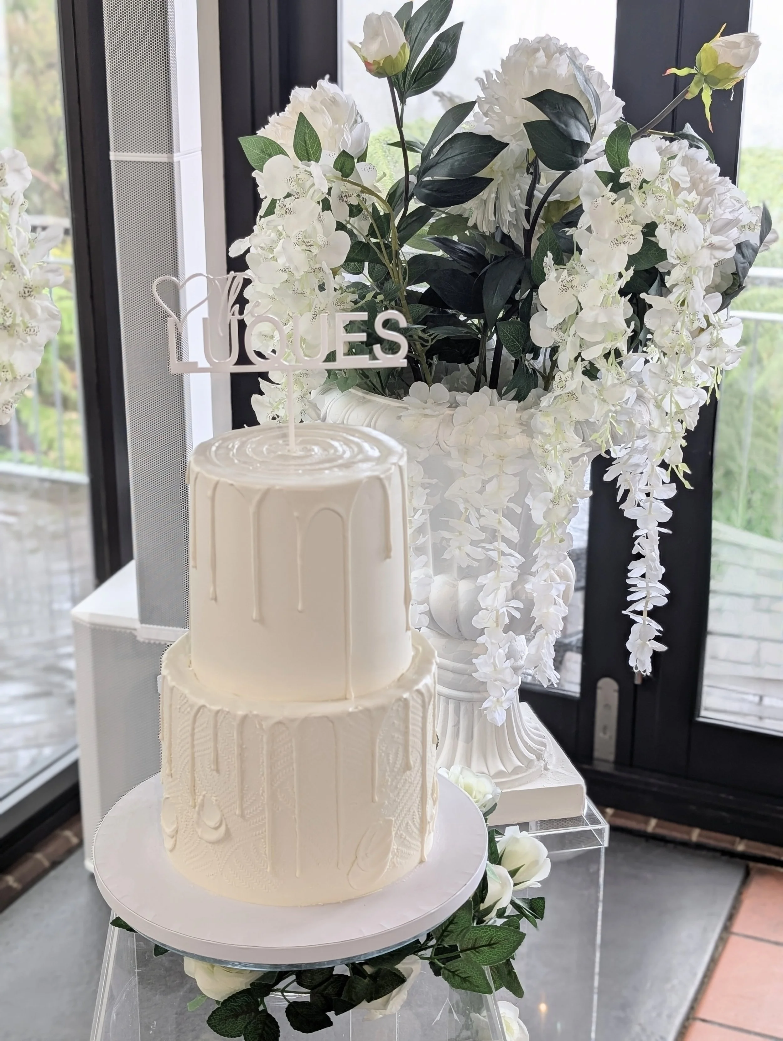 A two-tier white wedding cake with a 'LOVE LOVES' cake topper, decorated with white drips and textured patterns, placed on a stand with white roses and greenery, next to a large floral arrangement of white hydrangeas and other flowers in a white urn,
