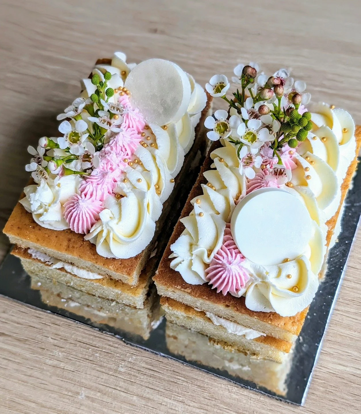 Decorative layered cream cakes with white and pink frosting, topped with white flowers, greenery, gold beads, and round edible decorations, on a reflective silver tray.