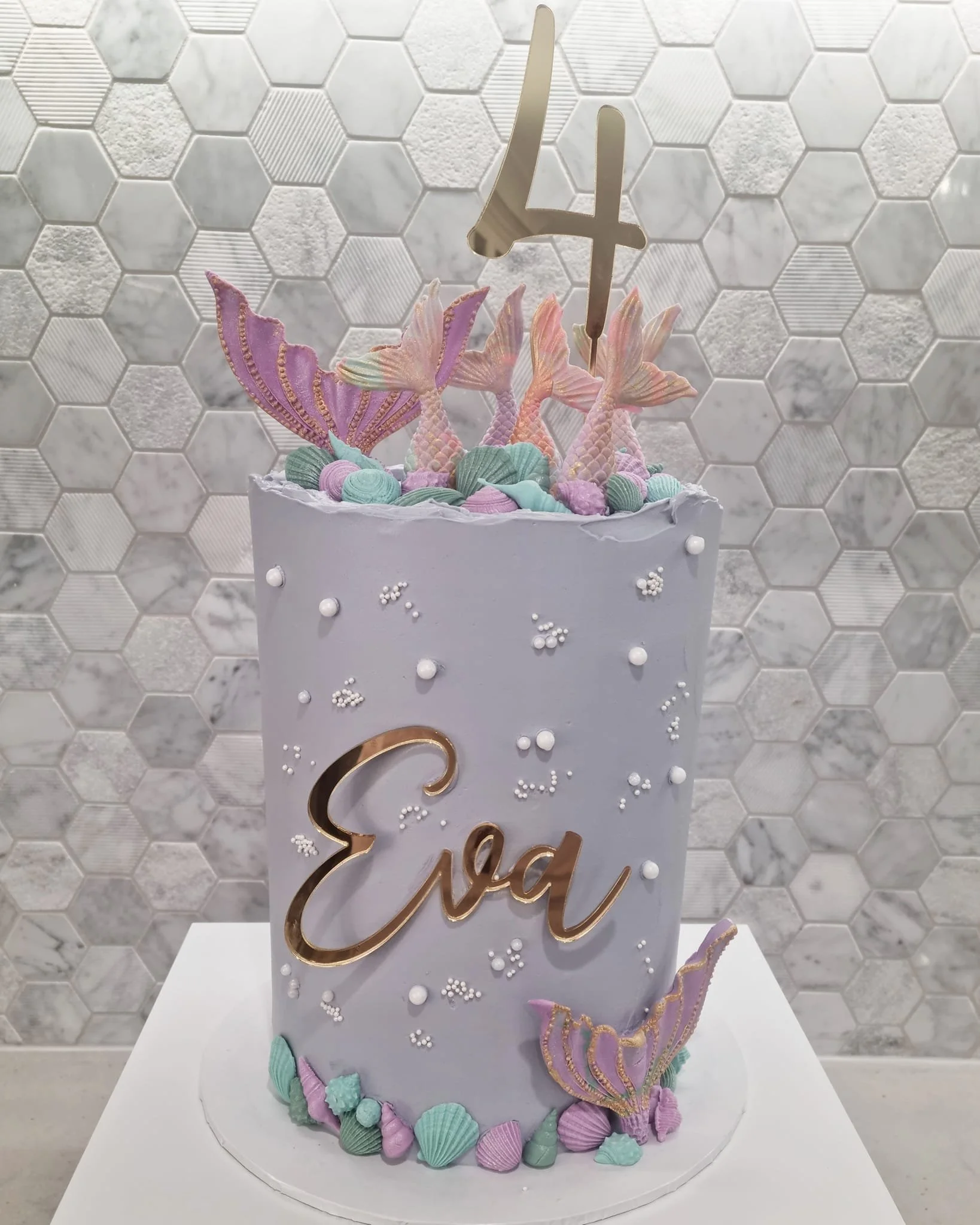 A pastel-colored birthday cake decorated with mermaid and ocean-themed accents, including sea shells, fins, and tails, with a gold "Eva" topper and a large gold number four on top.