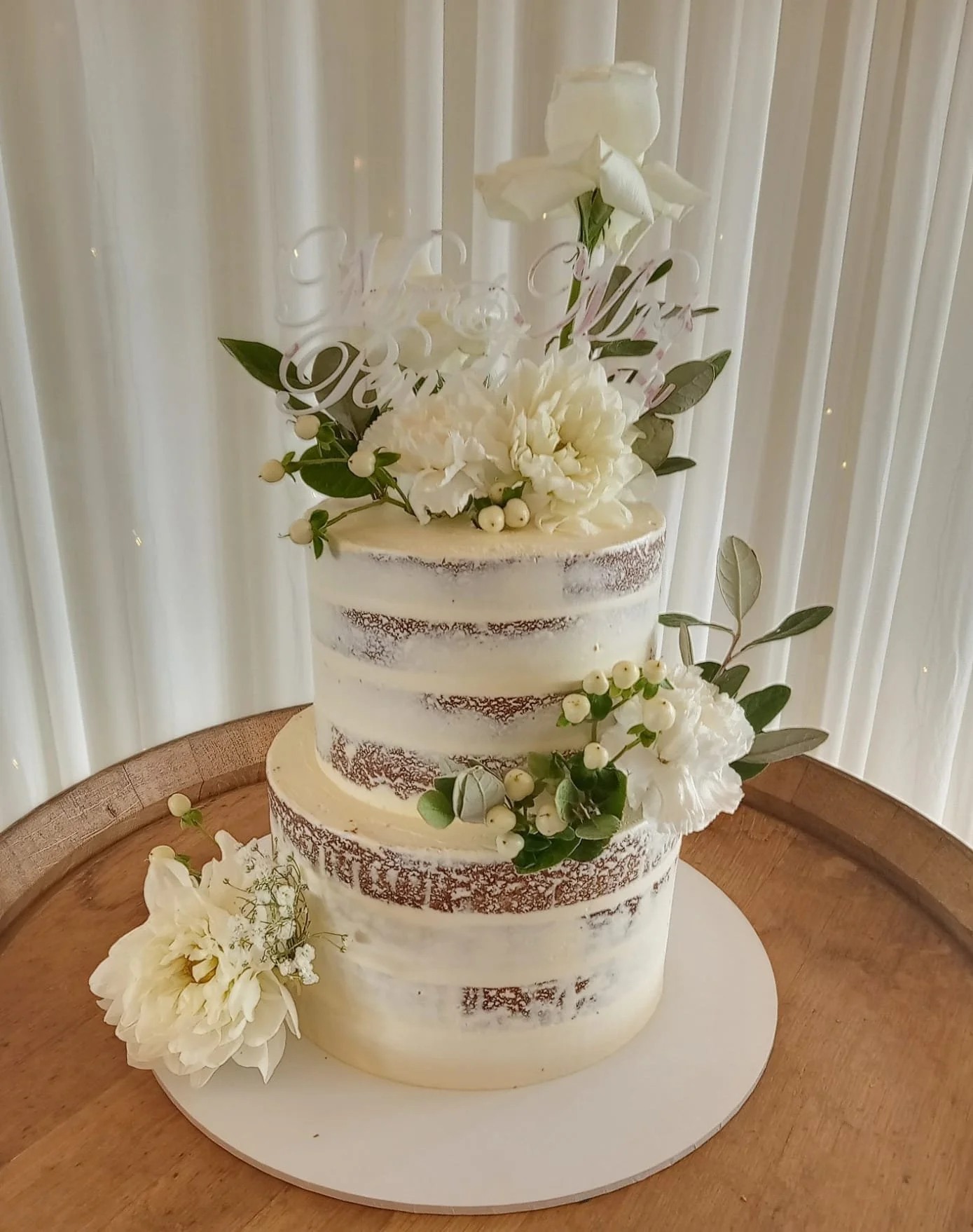 White semi-naked three-tier wedding cake decorated with white flowers and greenery, placed on a wooden table with a light-colored background.