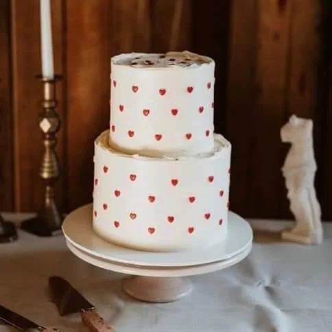 Two-tiered white cake with red heart decorations on a white cake stand, set on a table with a brass candlestick and ceramic horse figurine in the background.