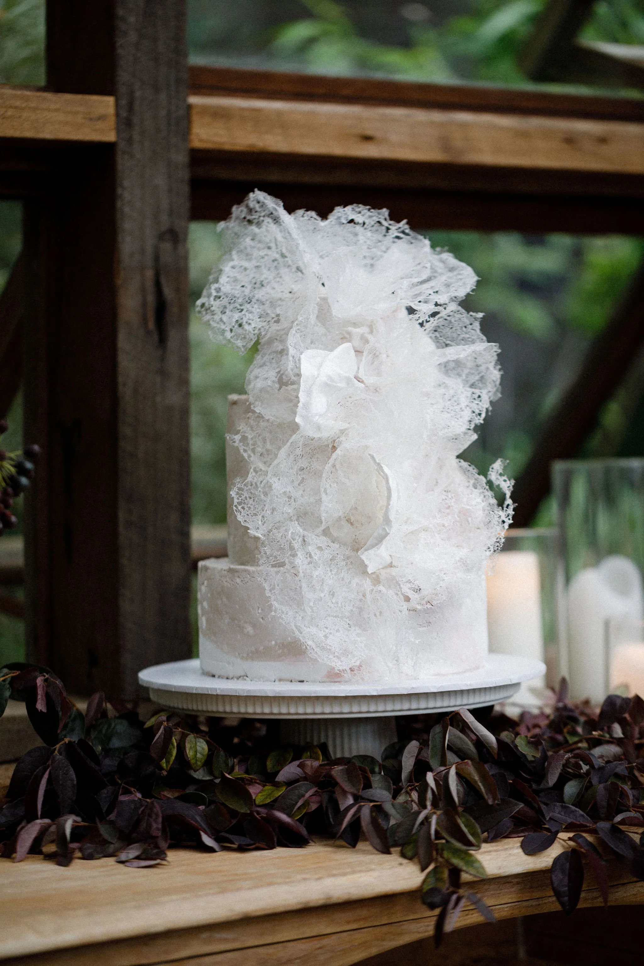 Elegant white cake with decorative lace-like sugar sculptures on top, placed on a white cake stand, surrounded by dark green and purple foliage, with candles in glass holders in the background.