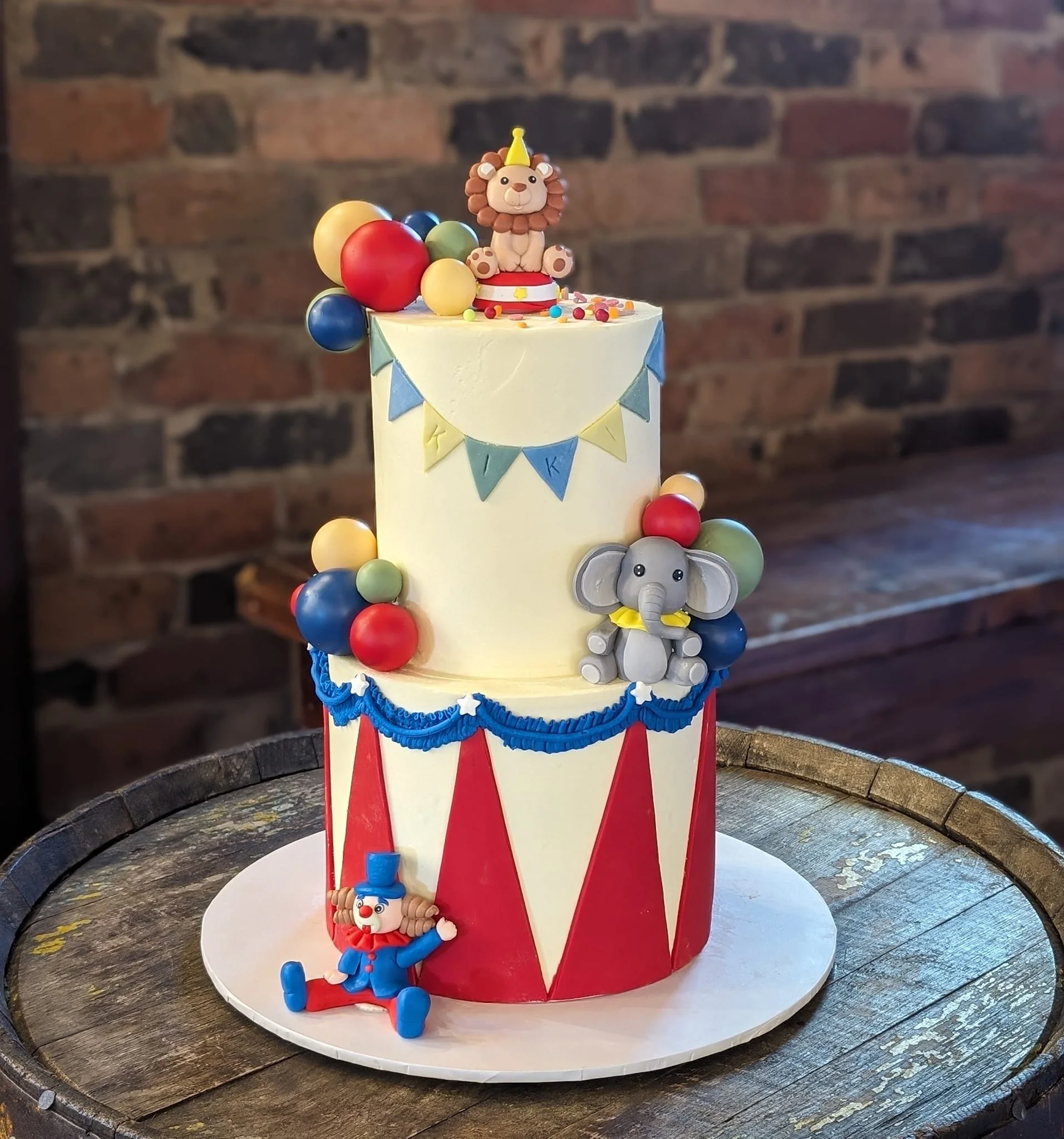 Two-tier circus-themed birthday cake with clown figure on top, colorful balloons, and elephant and lion decorations, on a wooden table against a brick wall.
