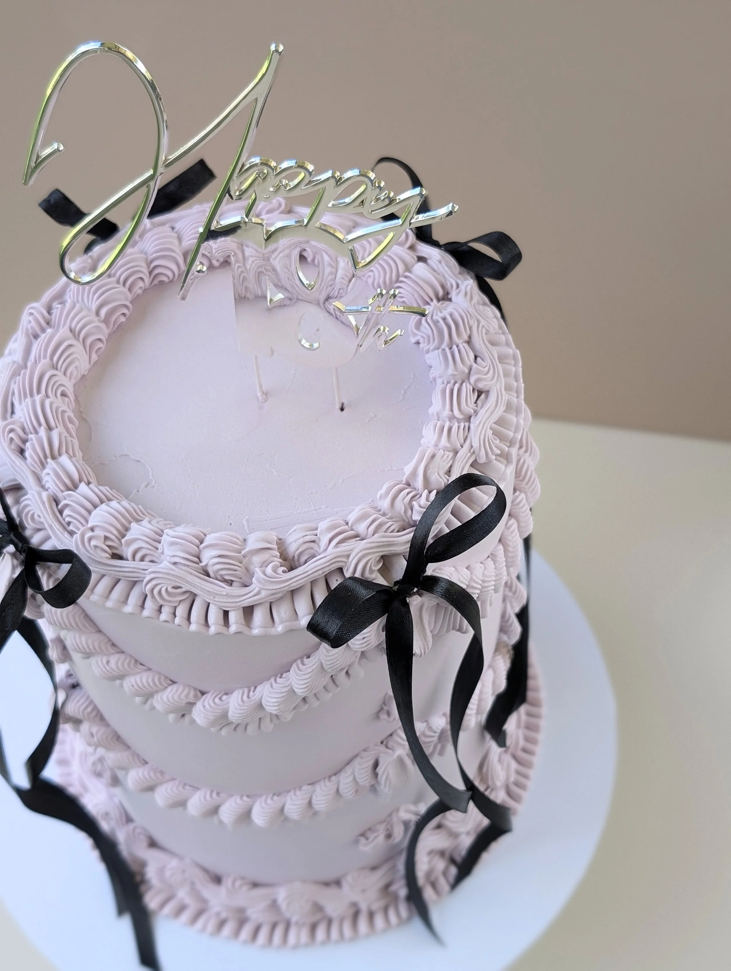 A tall, layered birthday cake decorated with pink frosting, black ribbon bows, and a gold "Happy Birthday" cake topper.