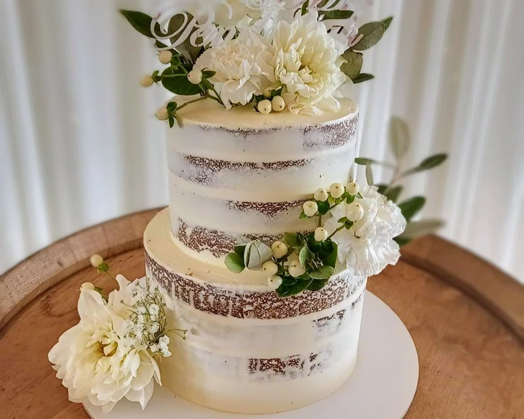 Two-tier semi-naked wedding cake decorated with white flowers and greenery, placed on a wooden surface.