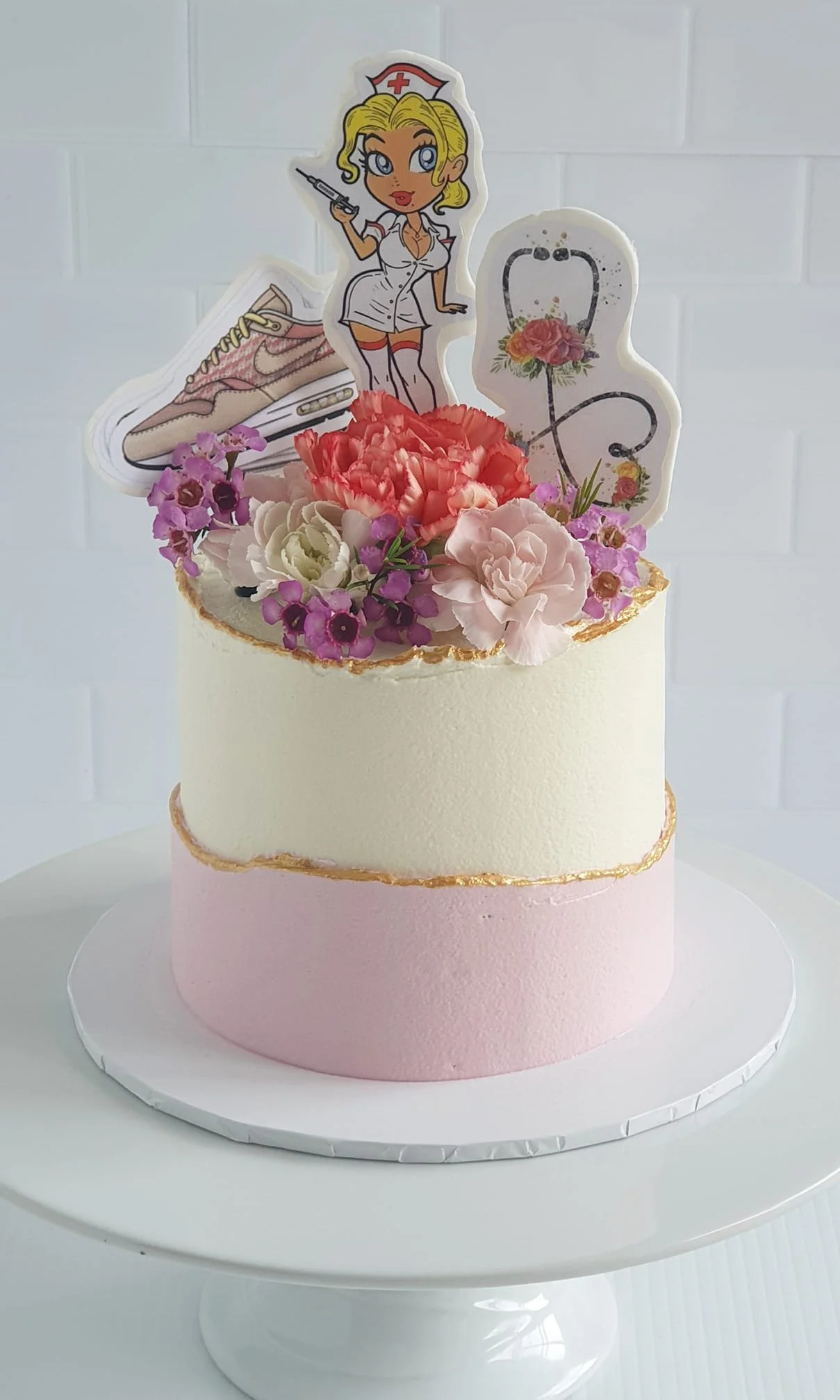 Two-tiered cake with pink bottom layer and white top layer, decorated with pink and white flowers, and cartoon nurse and medical-related toppers with flowers and stethoscope.