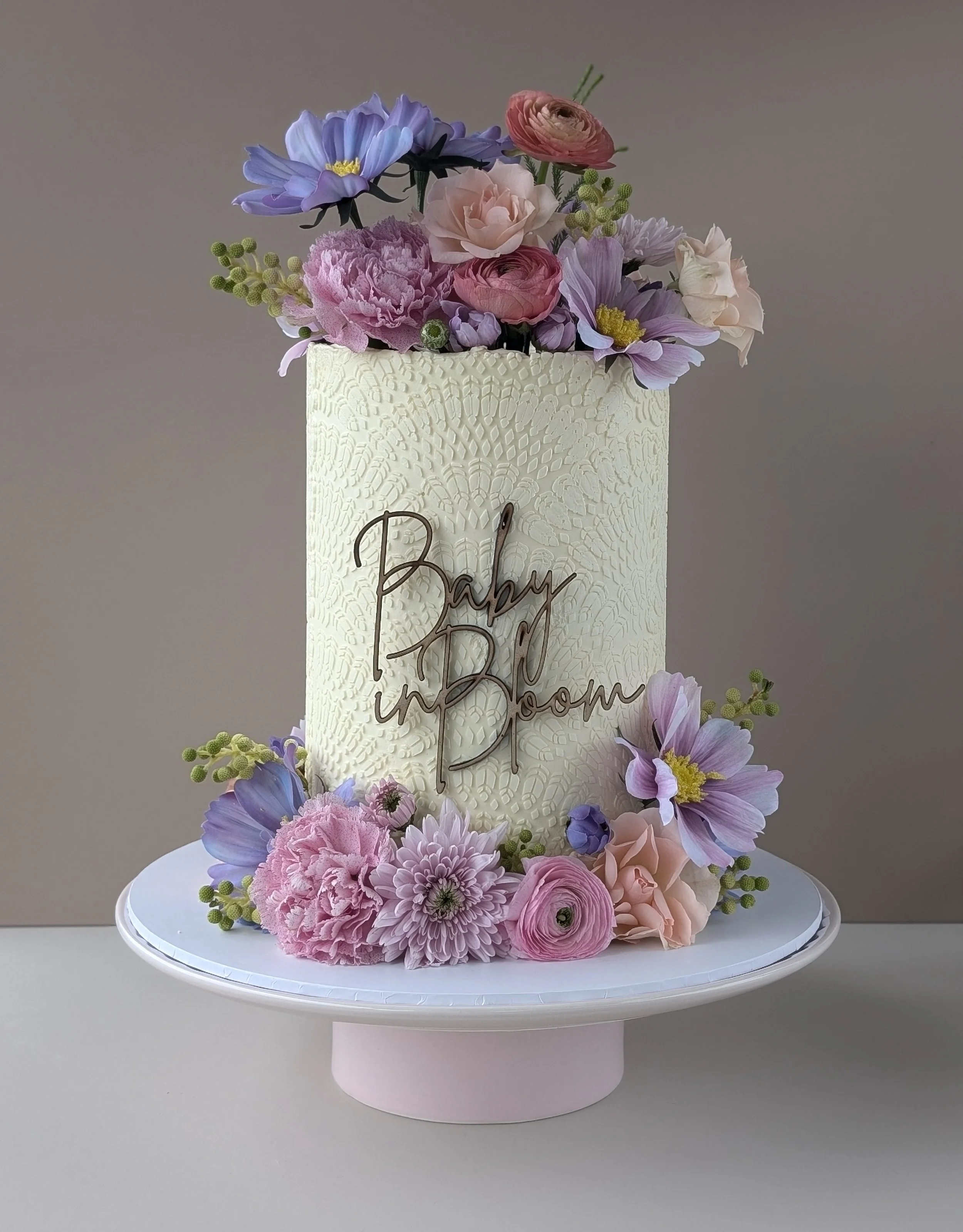 A tall white cake decorated with pastel-colored flowers and a sign that reads 'Baby in Bloom'