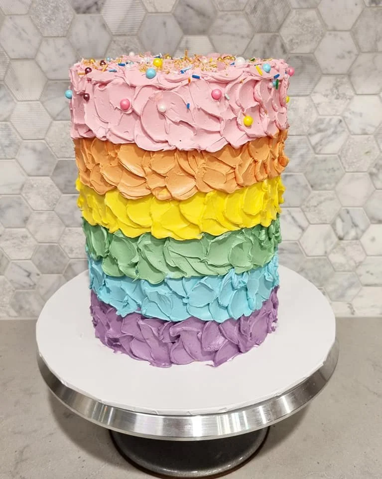 A six-layer rainbow cake with pink, orange, yellow, green, blue, and purple frosting, topped with colorful sprinkles and small round candies.