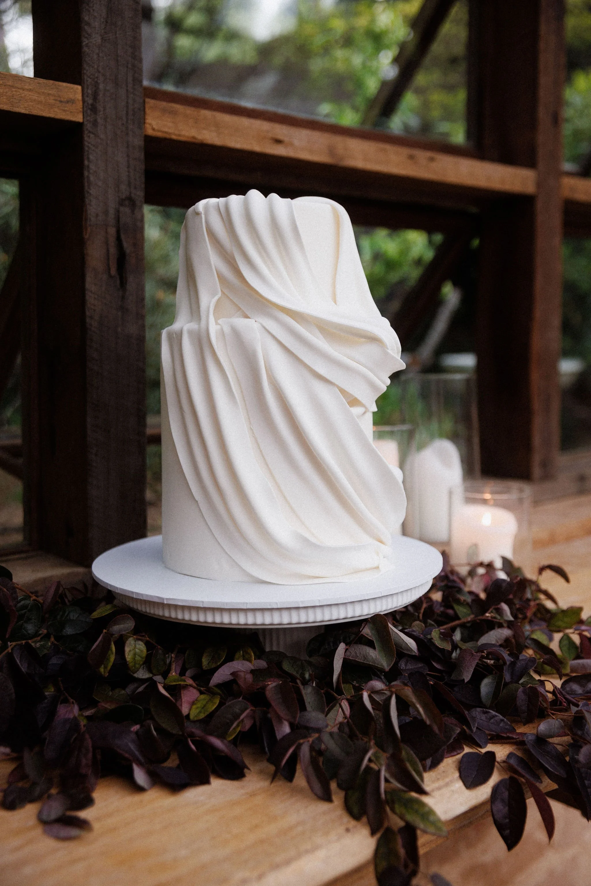 Sculpture resembling a human face with draped fabric features, white in color, on a round pedestal. Decorated with dark leaves and surrounded by candles in glass holders.