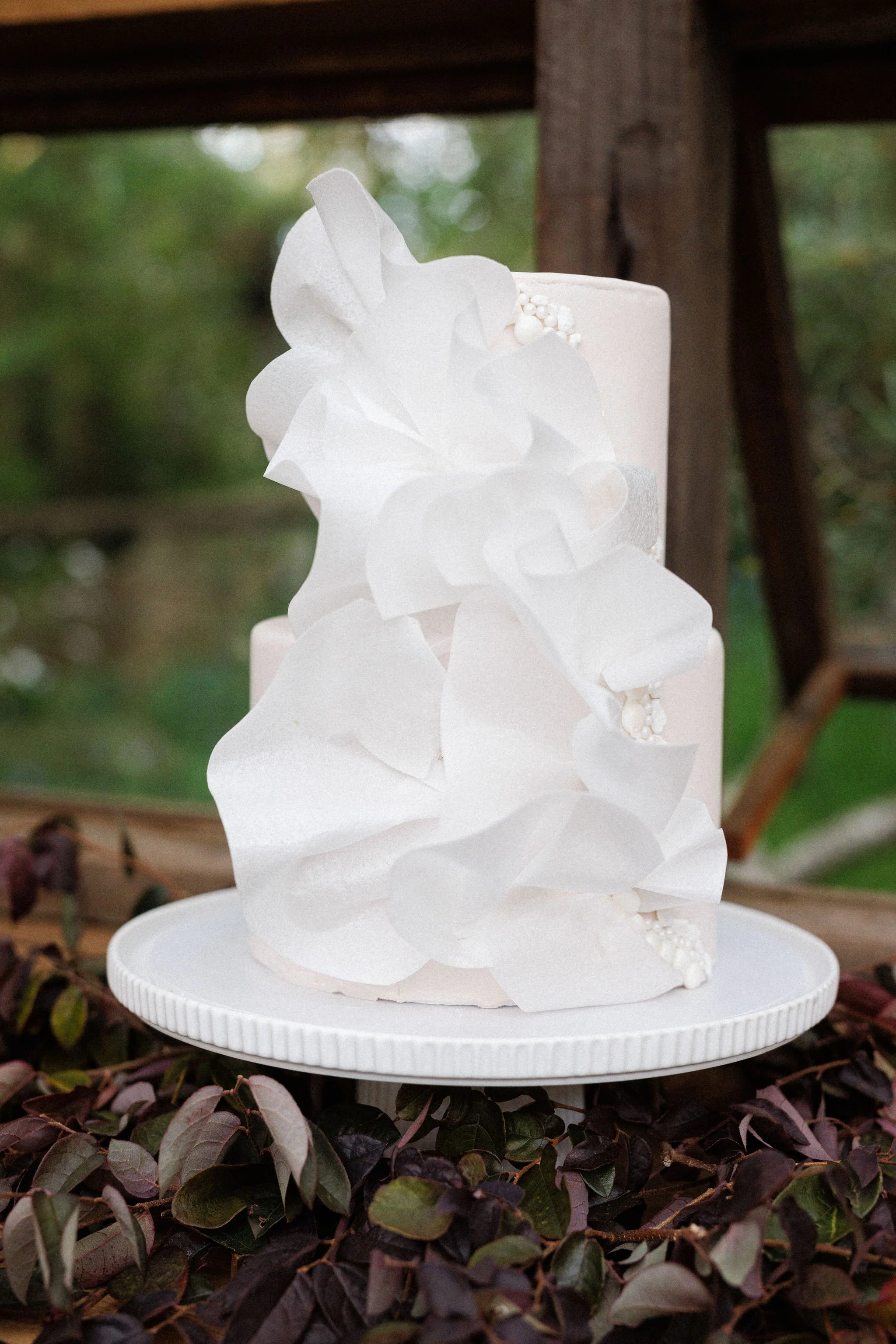 A two-tier white wedding cake decorated with white paper or fabric flowers and small white beads, placed on a white round cake stand outdoors surrounded by purple and green foliage.