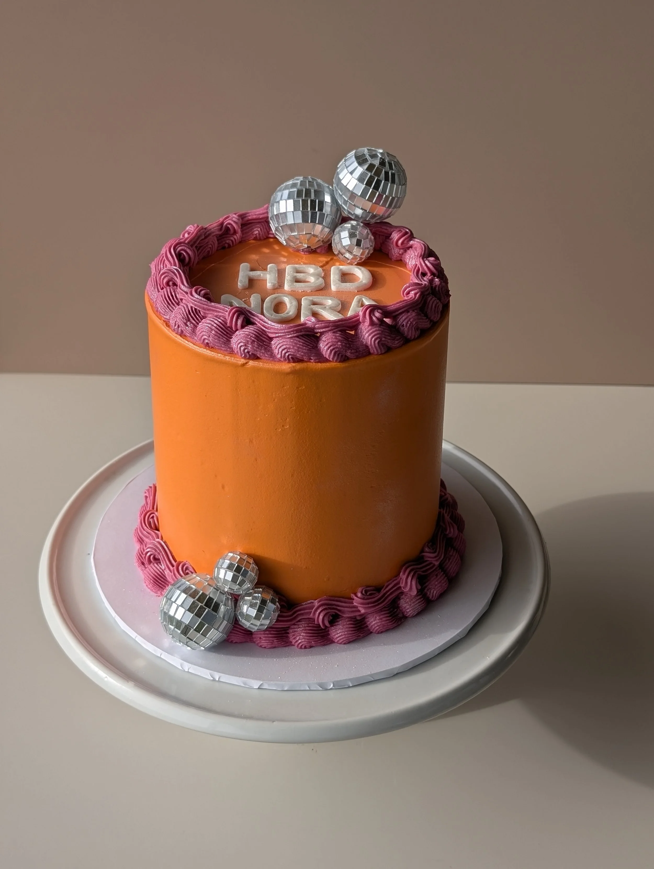 Celebration cake with orange icing, pink piping, disco ball decorations, and the words 'HBD' on top, on a white plate.