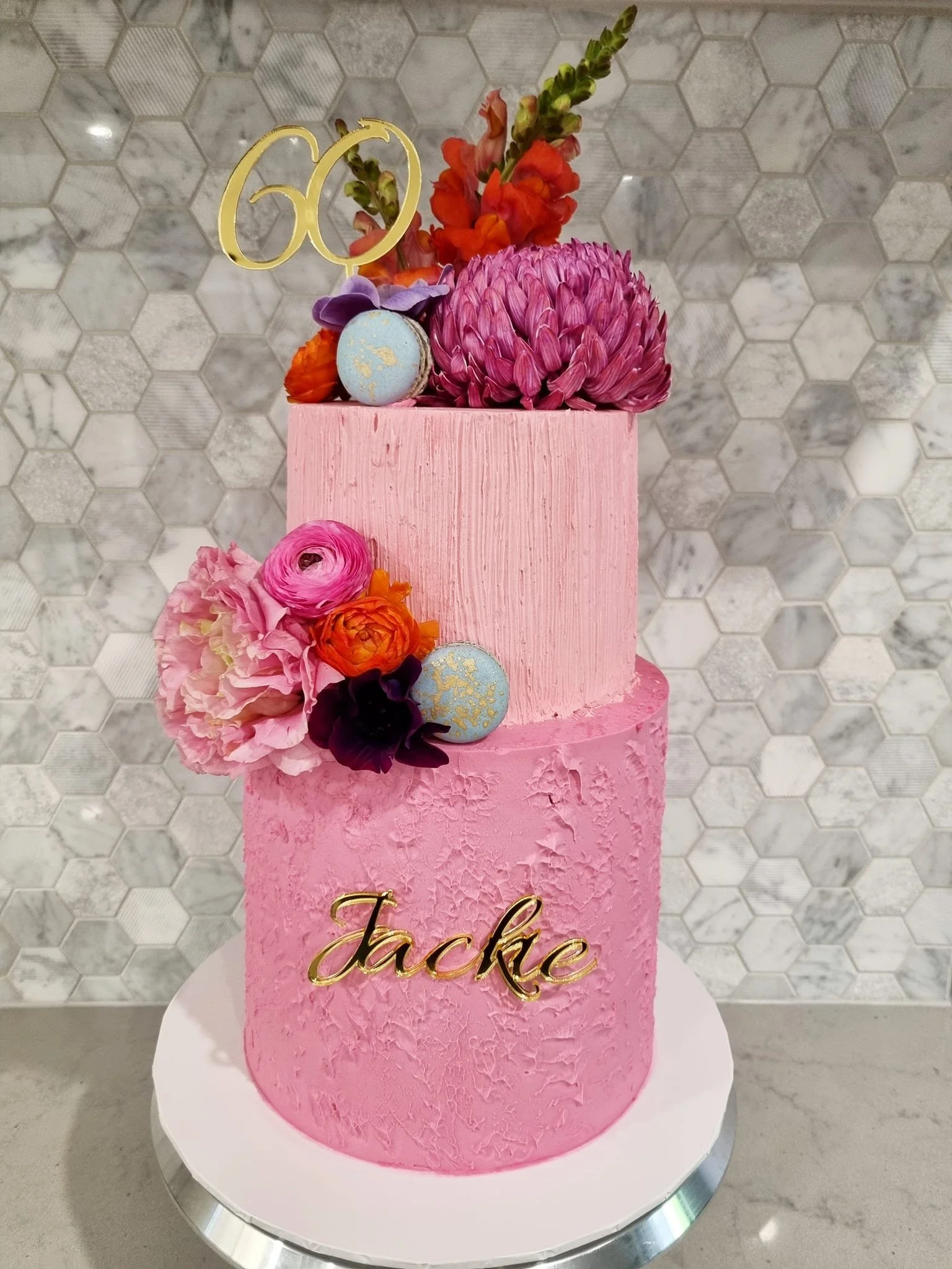 Pink two-tiered cake decorated with flowers, macarons, and a gold 